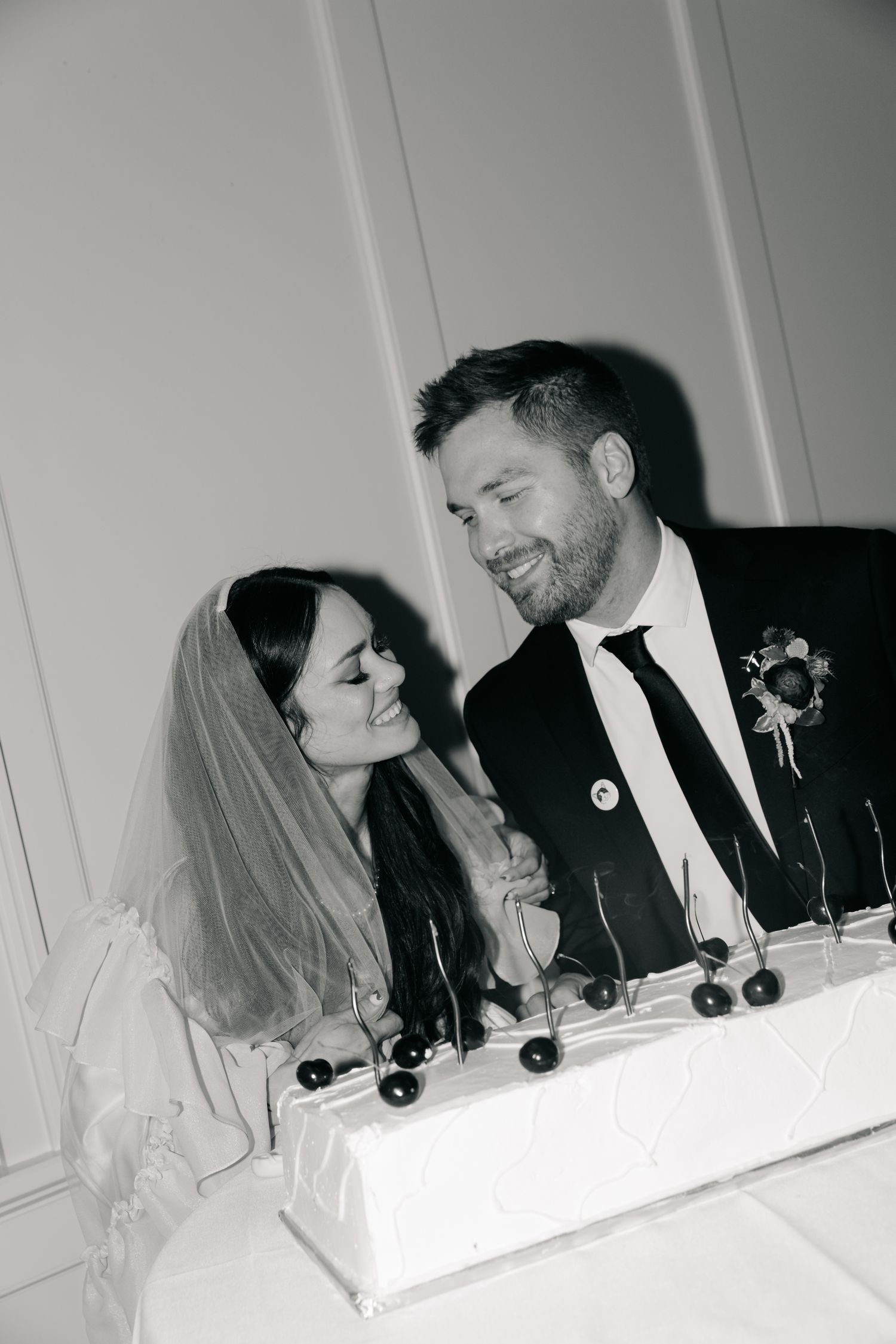 Black and white photo of a smiling bride and groom cutting their wedding cake together at their reception.