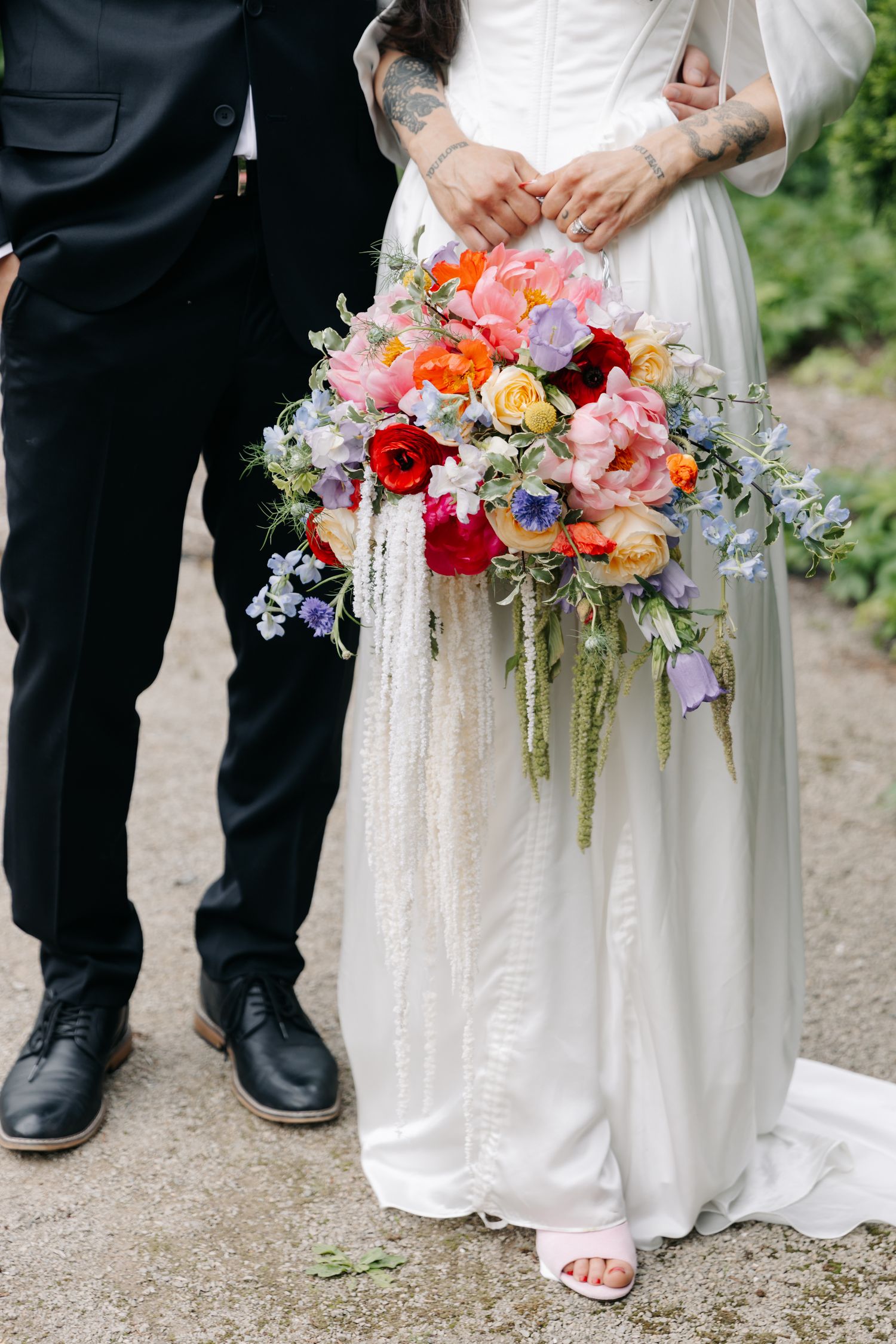 Bride in white gown holding colorful wedding bouquet with pink, red, and purple flowers beside groom in black suit.