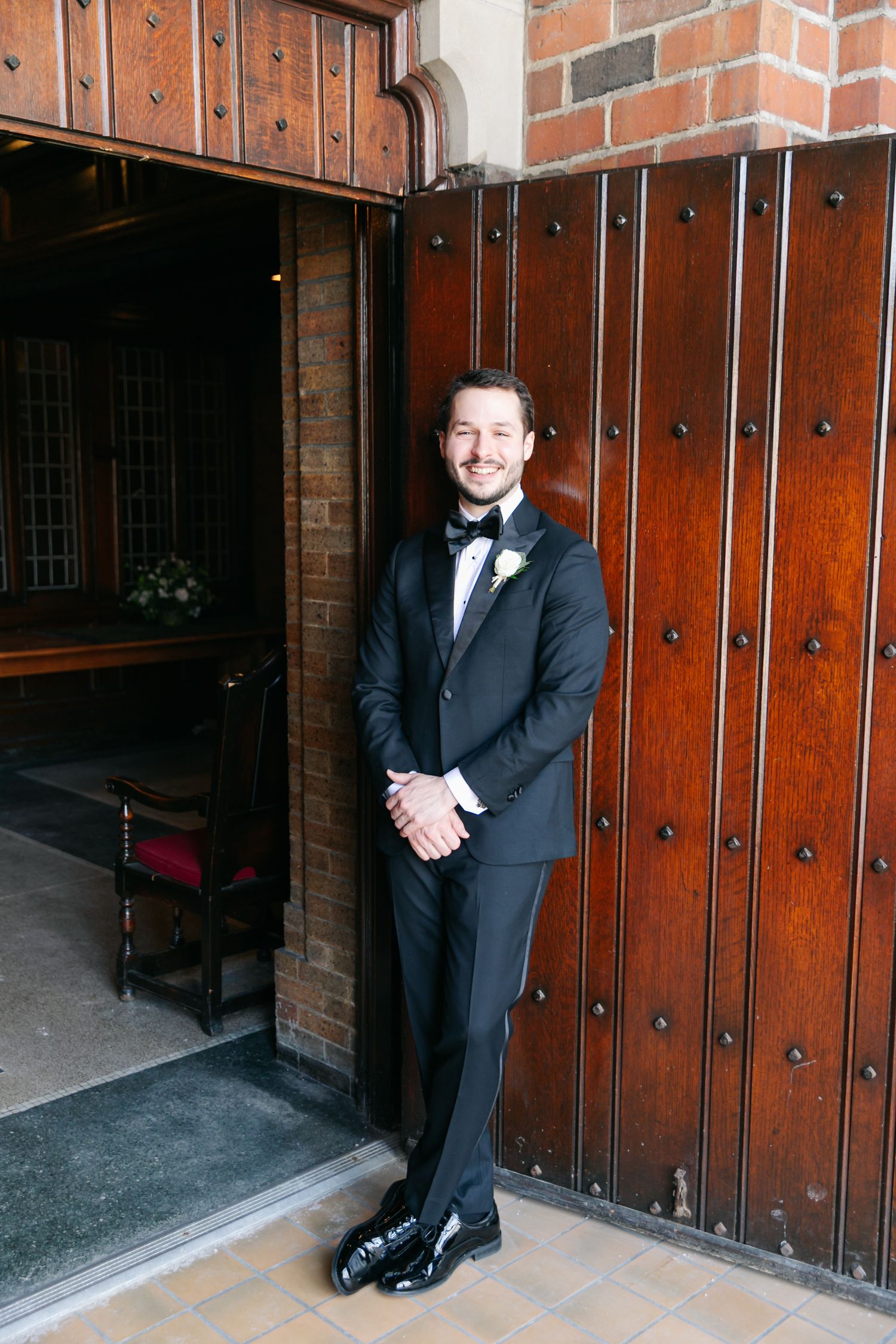 Smiling groom in black tuxedo with bow tie and boutonniere standing by ornate wooden church doors.