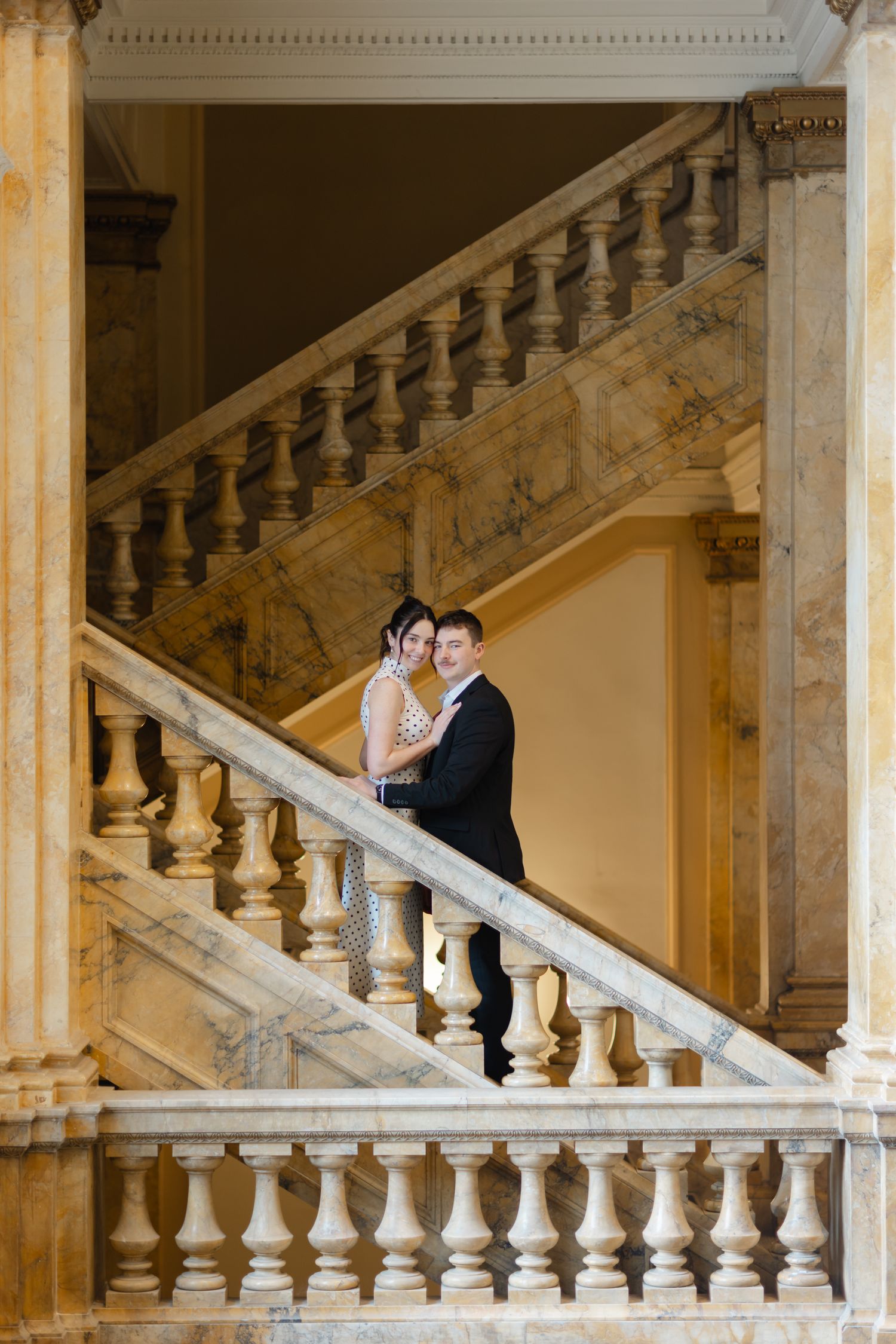 Couple embracing on ornate marble staircase with elegant balustrade in a grand classical building.