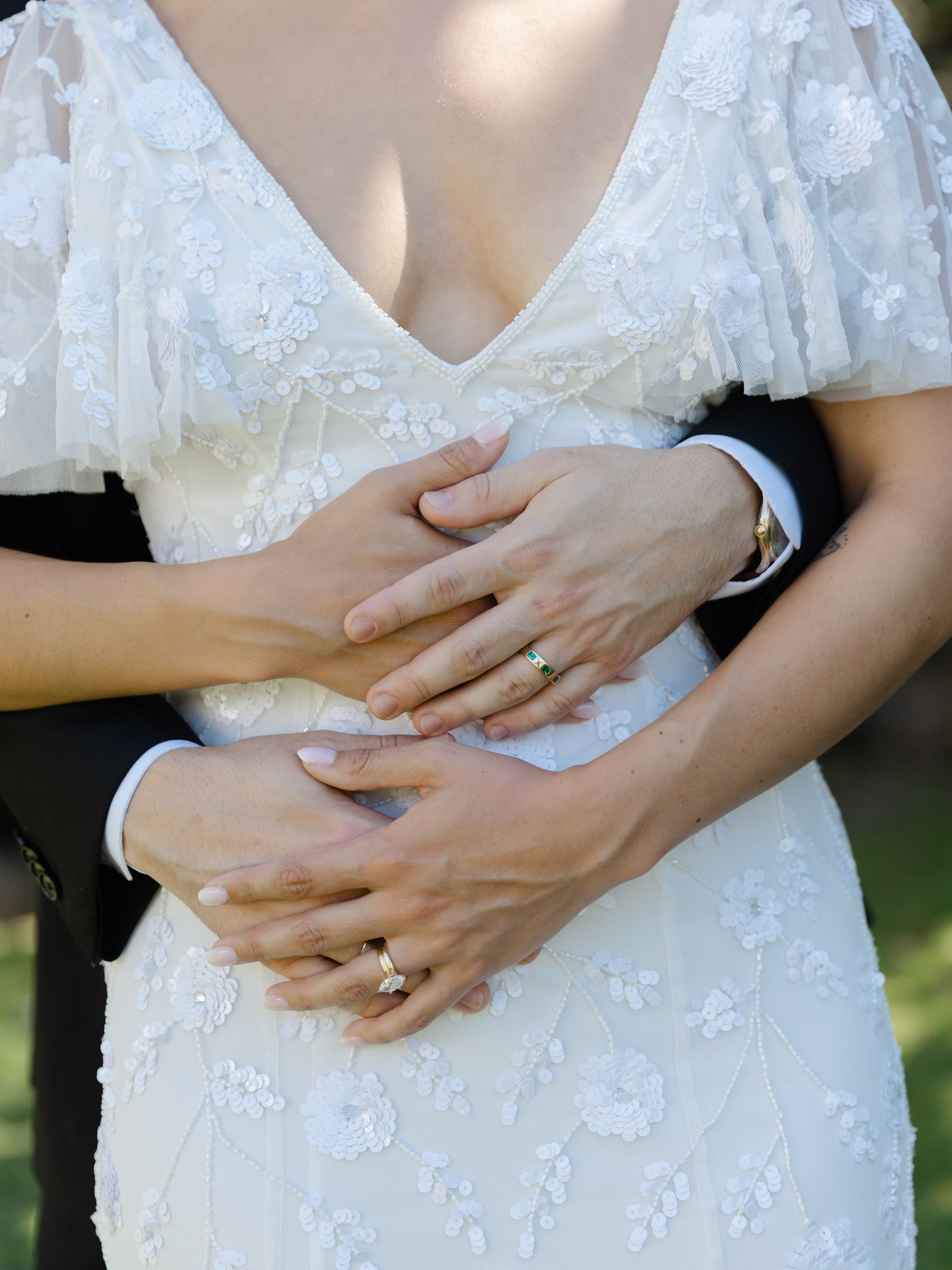Bride and groom holding hands at waist, showing wedding rings, bride in white floral lace dress.