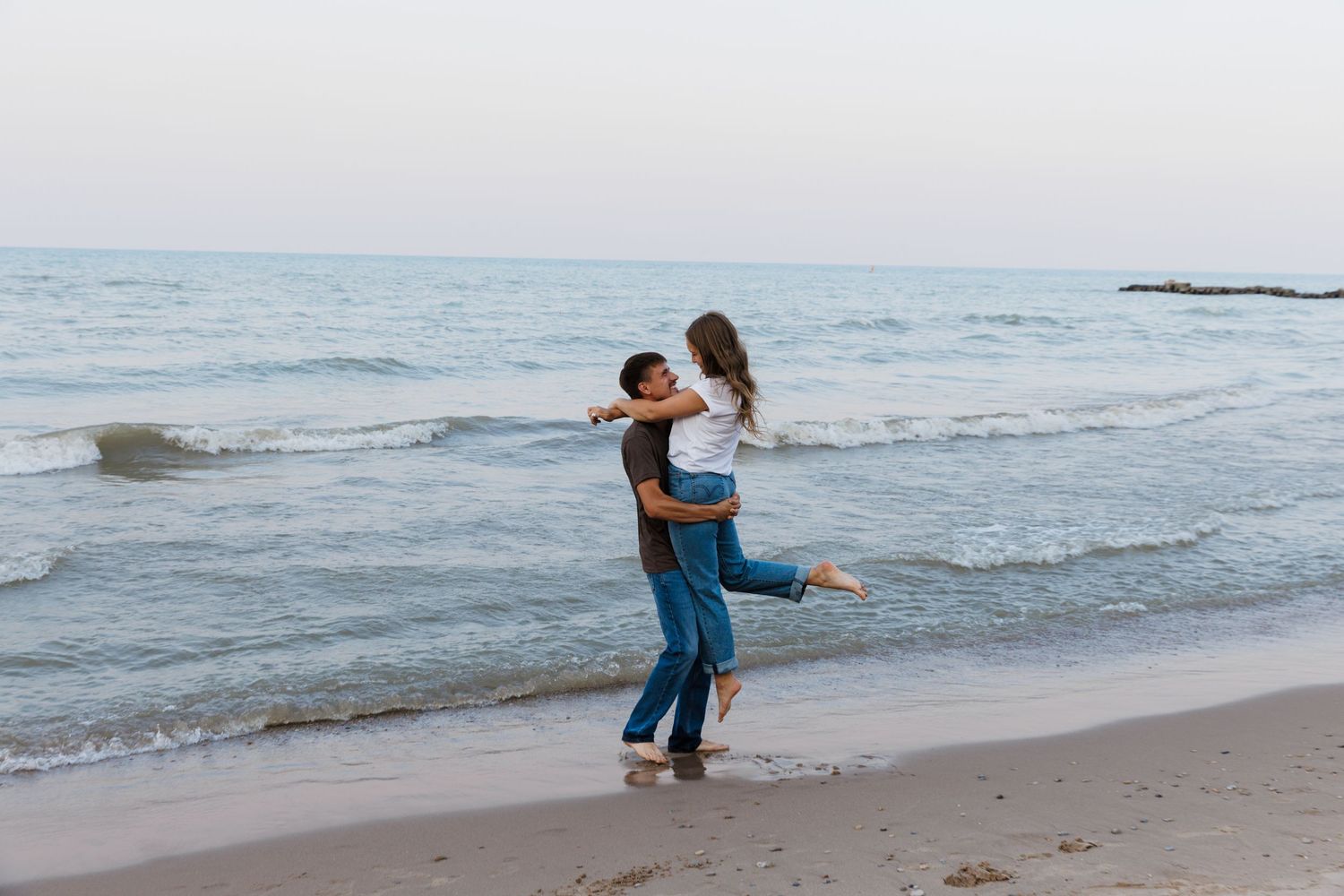 Romantic couple embracing on a sandy beach at dusk, man lifting woman near calm ocean waves.