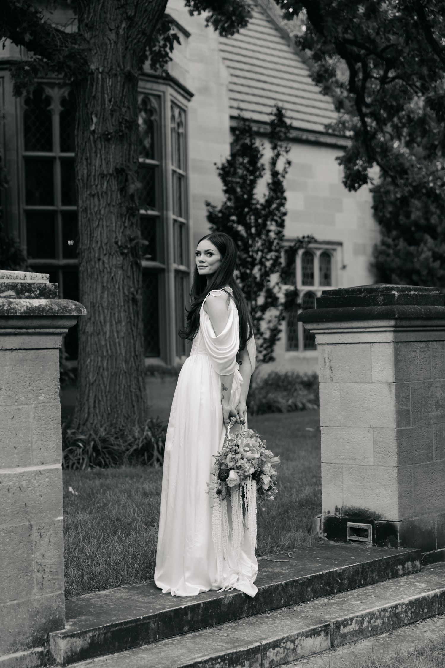 Bride in elegant white gown holding bouquet, posing on steps beside historic stone manor in black and white.