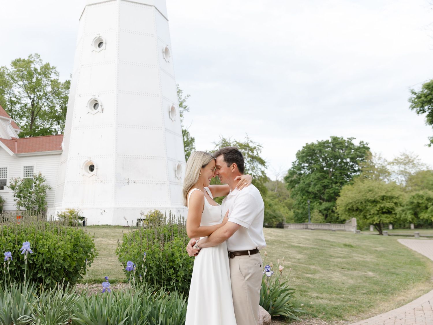 Couple sharing a romantic kiss in front of a white lighthouse surrounded by lush gardens and blooming irises.