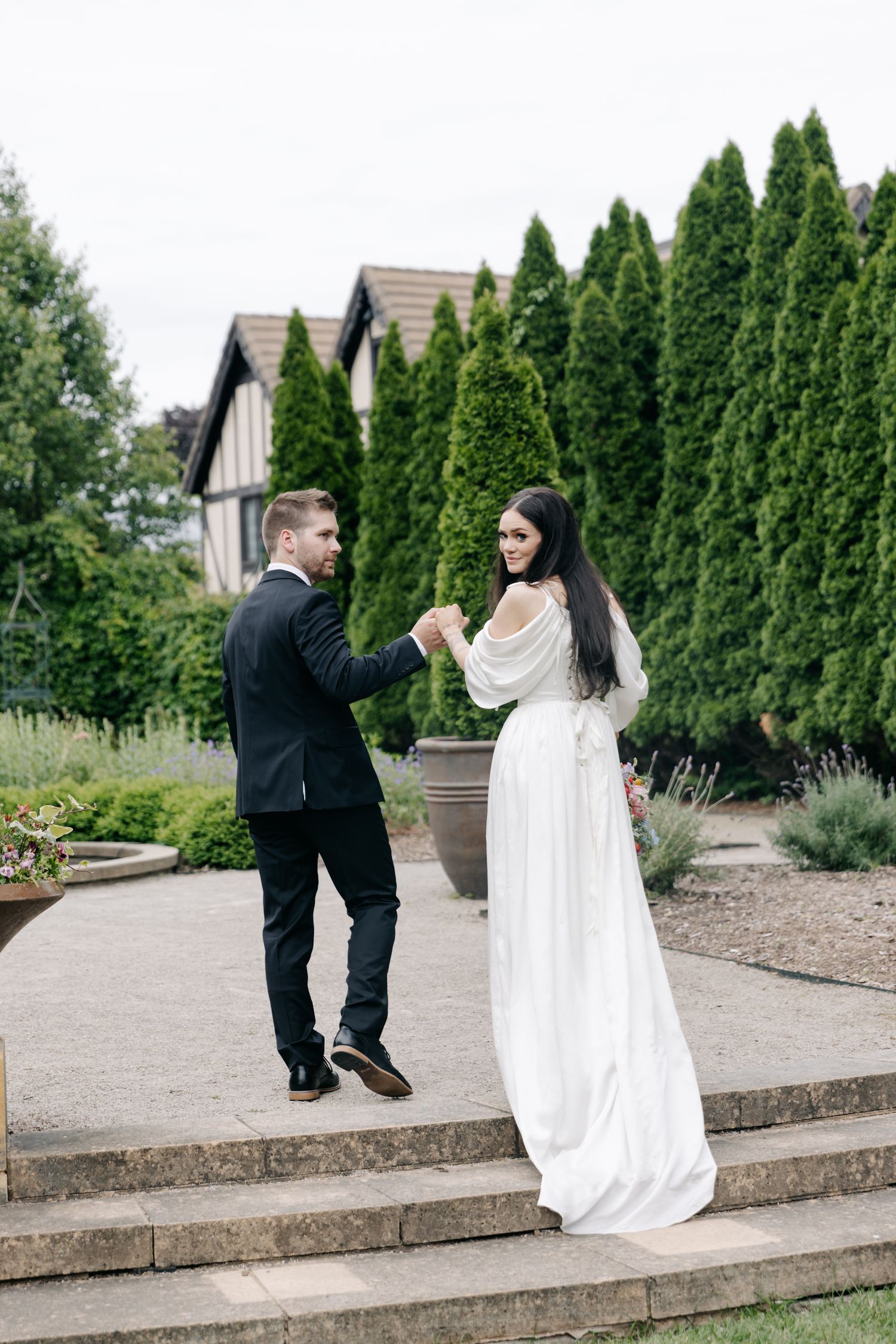 Bride in white gown and groom in black suit hold hands on garden steps with tall evergreen trees behind them.