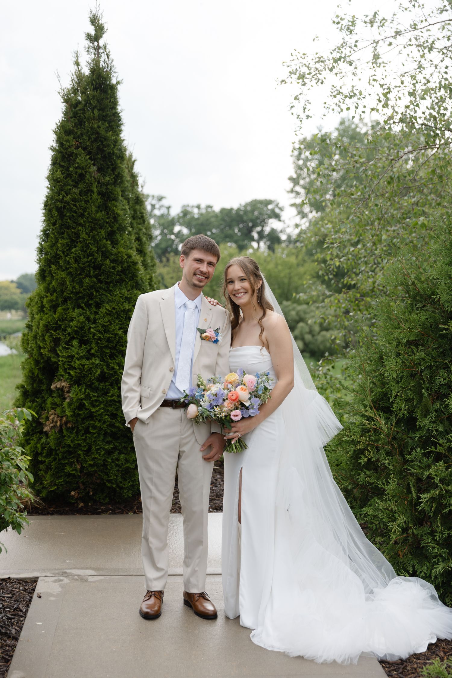 A bride and groom pose together outdoors on a garden path, she holds a colorful bouquet in a white gown, he wears a tan suit.