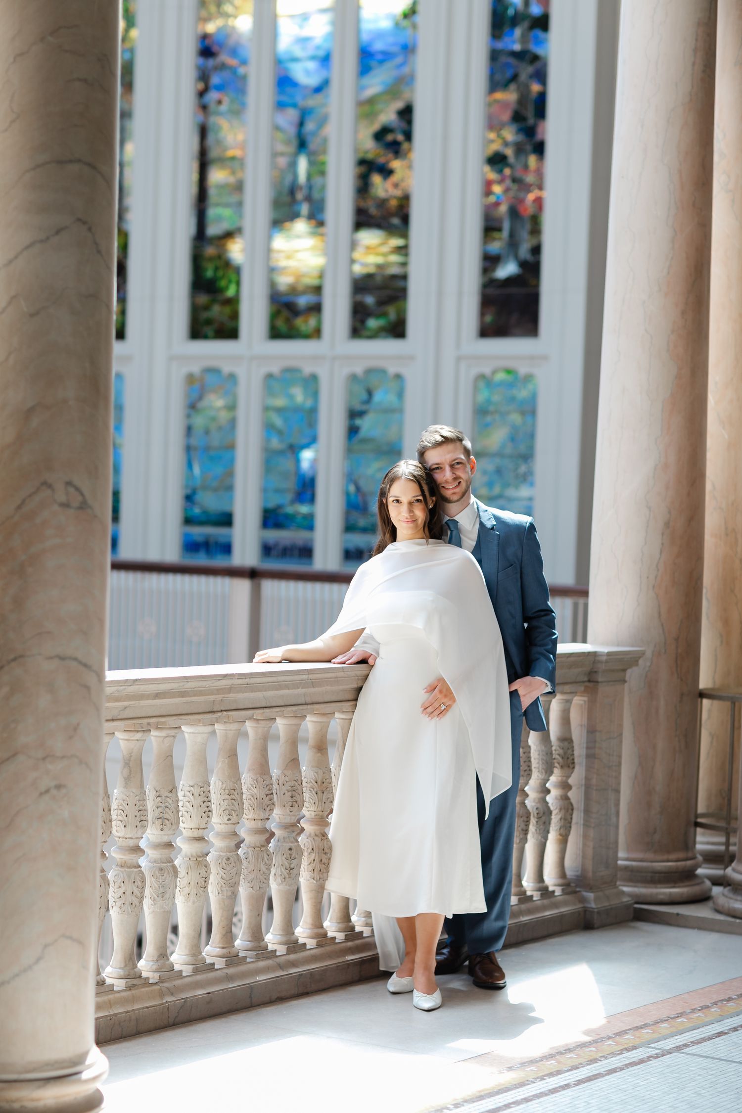 Elegant couple posing in white dress and blue suit before stained glass windows on marble balcony.