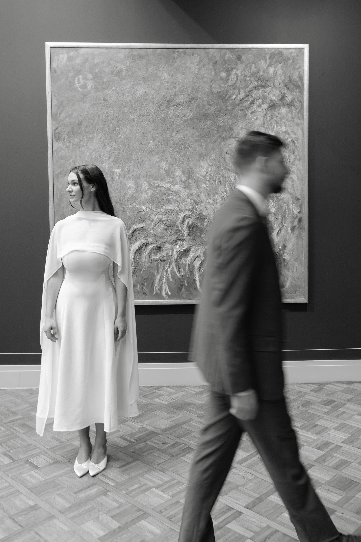 Black and white photo of a woman in white dress posing before a large painting in a museum, man in suit walking past.