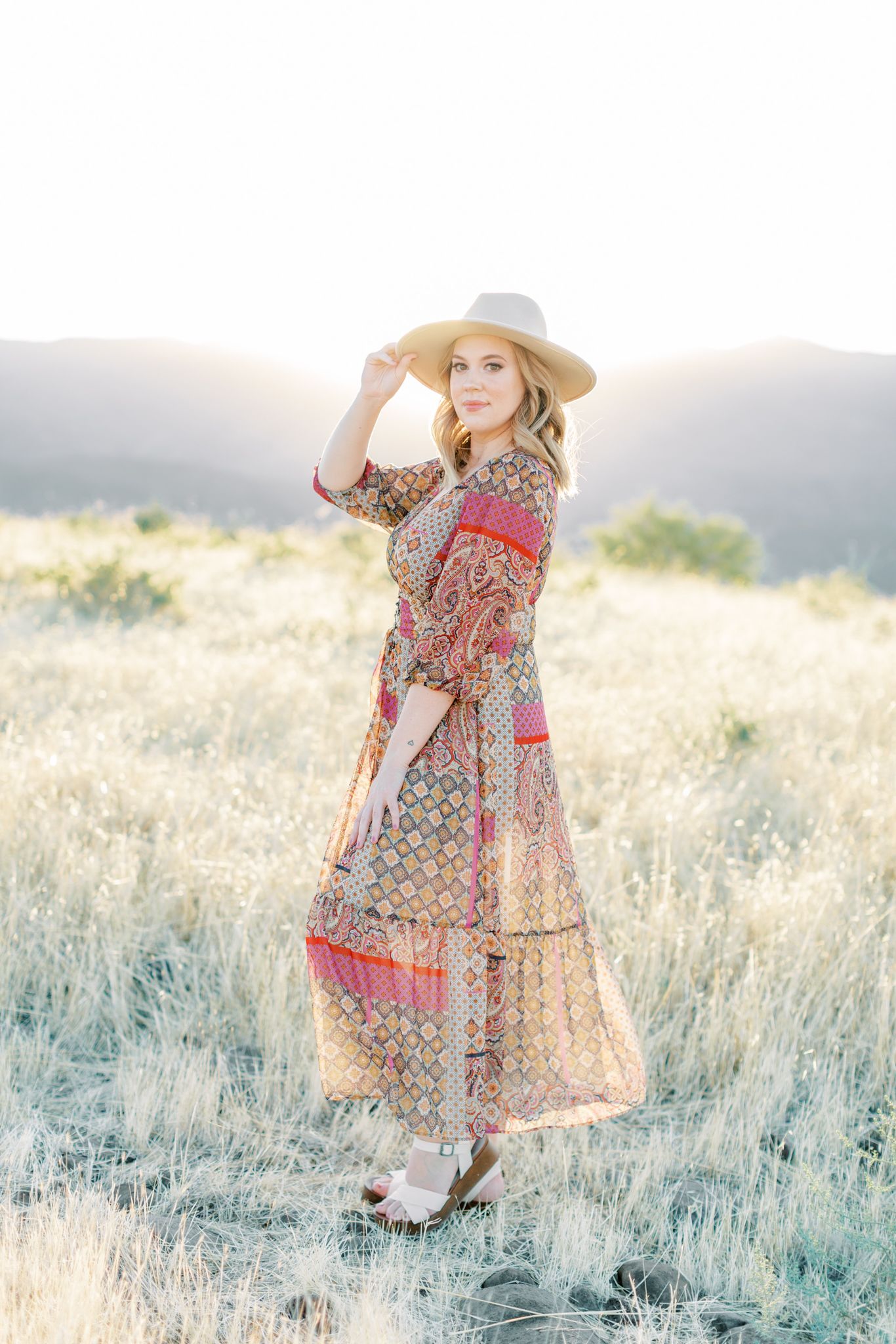 Woman in floral maxi dress poses in sunlit field wearing wide-brimmed hat.