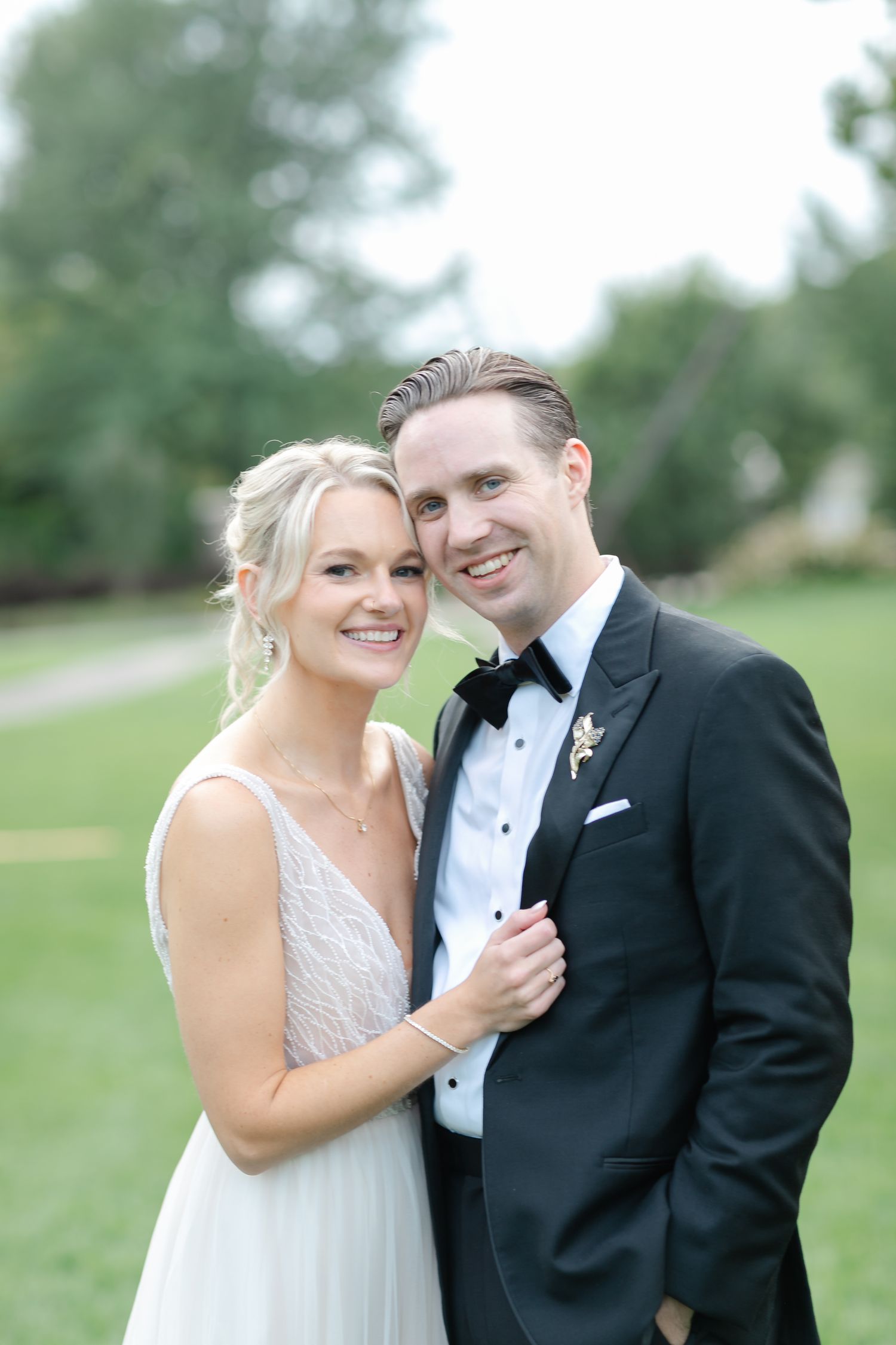 A smiling bride in a white gown and groom in a black tuxedo pose together outdoors on their wedding day.