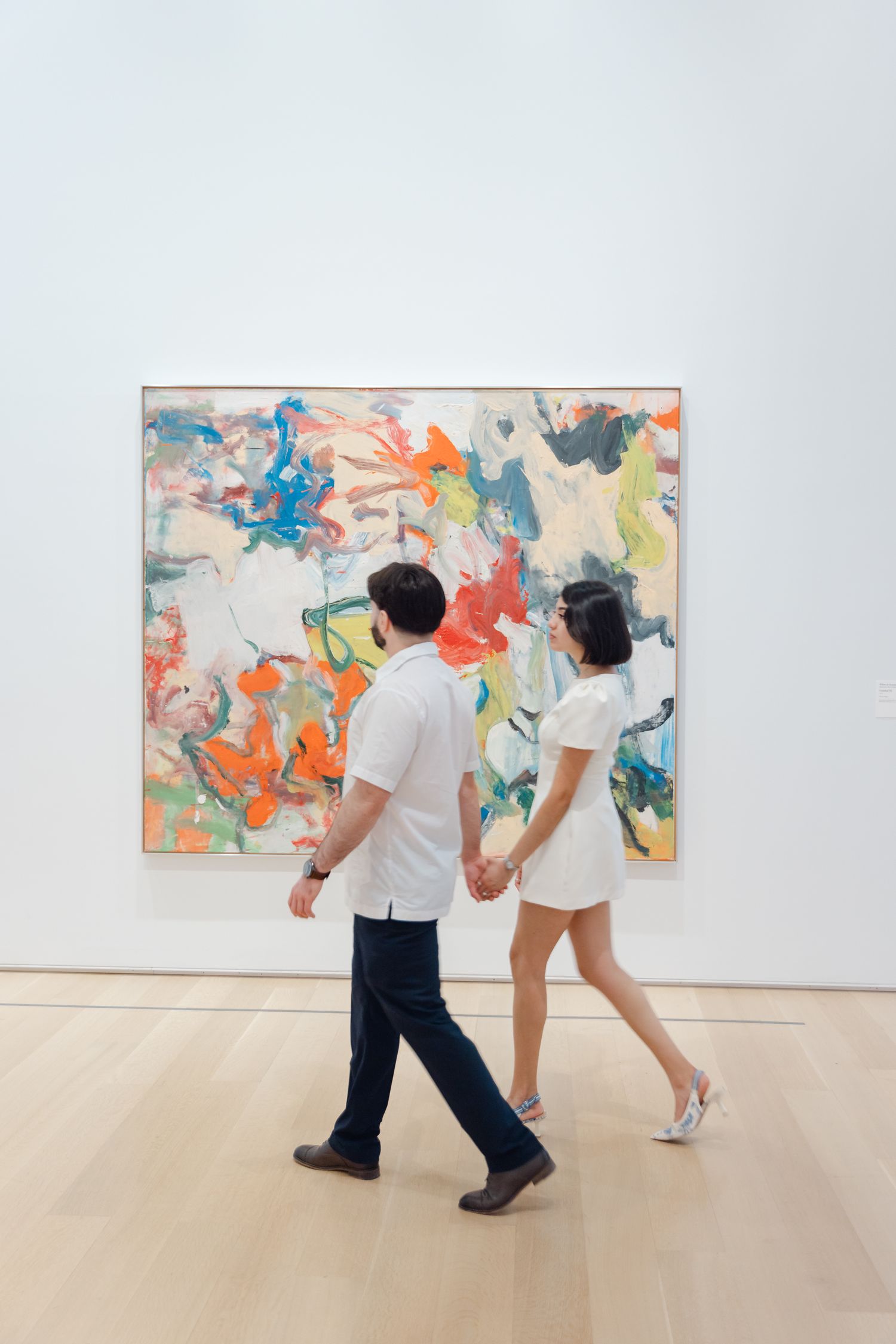 Couple holding hands walking past a large colorful abstract painting in a modern art gallery.