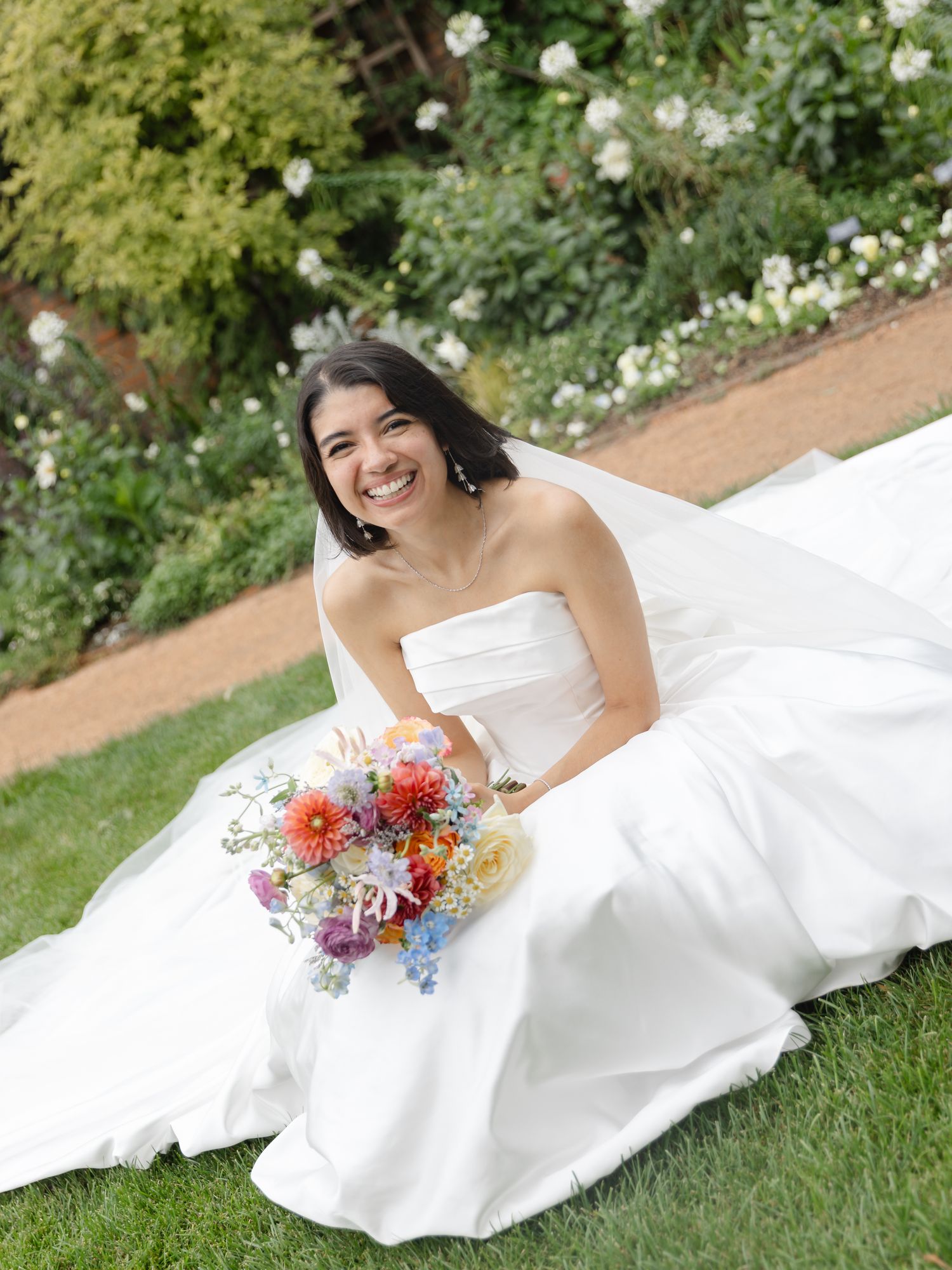 Smiling bride in a strapless white gown sits on grass holding a colorful bouquet, with lush garden greenery behind her.
