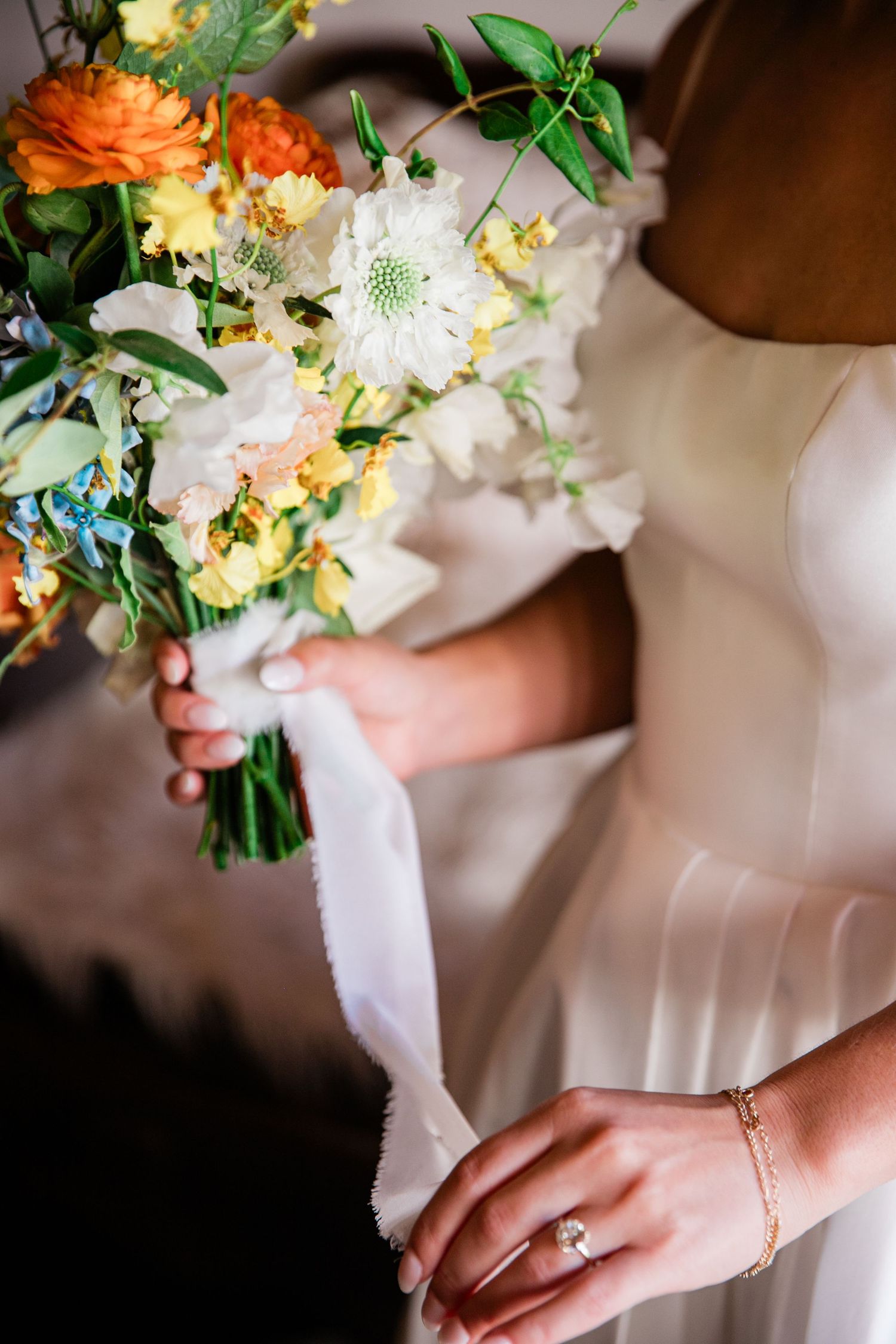 Close-up of a bridal bouquet with orange and white flowers, showing the bride's ring and bracelet details.