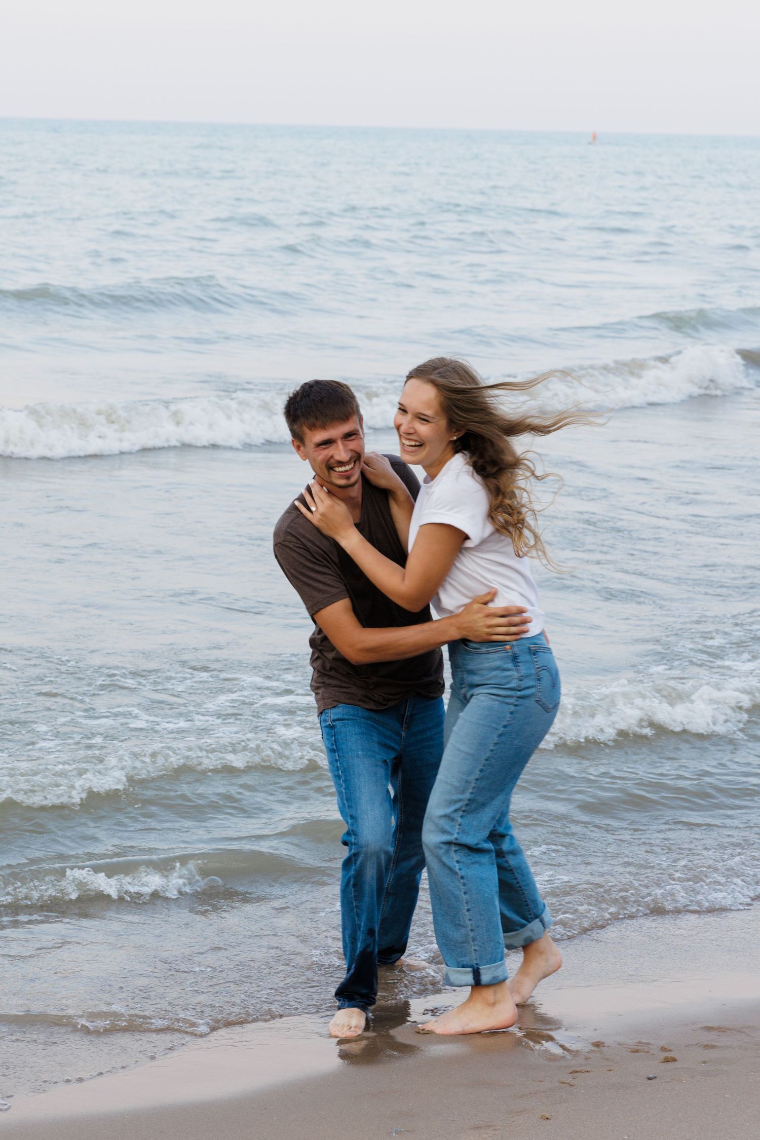 Happy couple embracing and laughing on a sandy beach with ocean waves in the background.