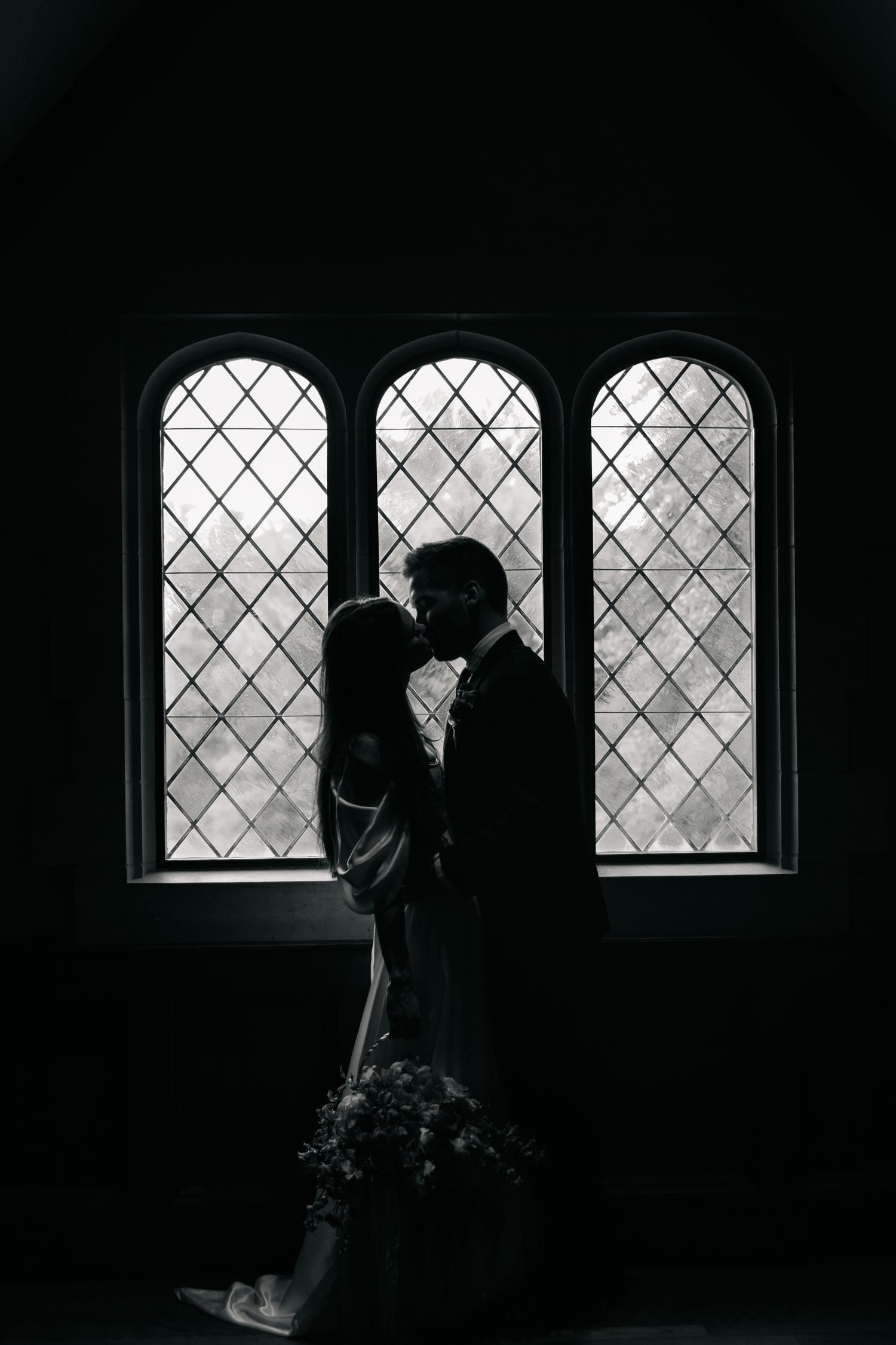 Silhouette of a couple kissing in front of arched stained glass windows in a dark romantic church setting.