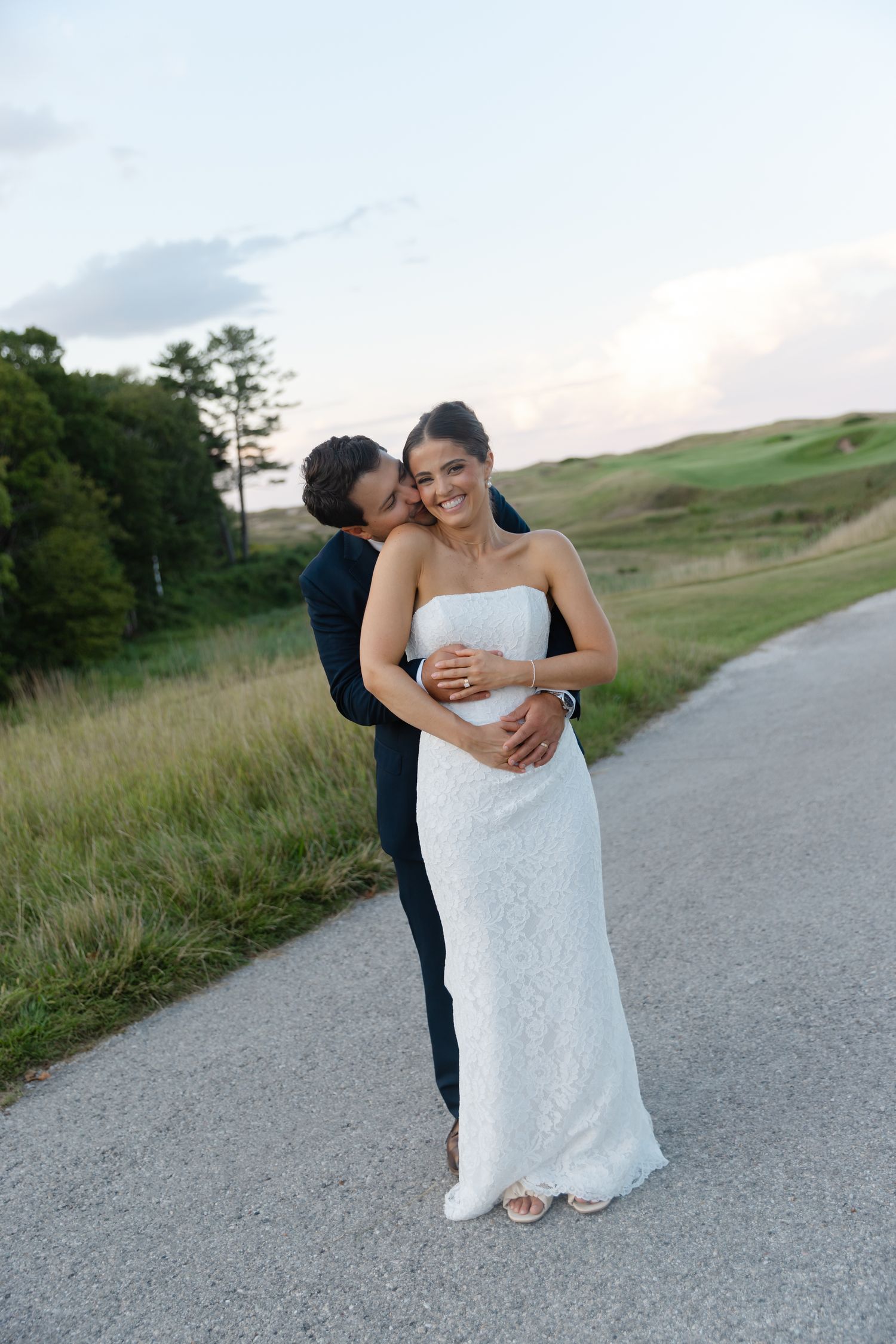 Bride in white lace gown embraced by groom in navy suit on a scenic road at dusk.