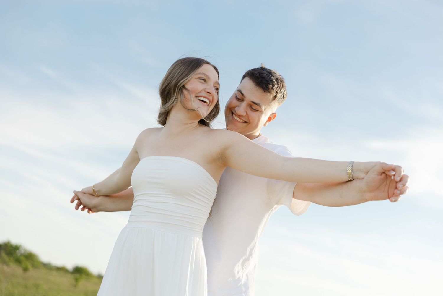 Happy couple in white attire embracing outdoors under a blue sky, smiling joyfully in a romantic wedding photo.