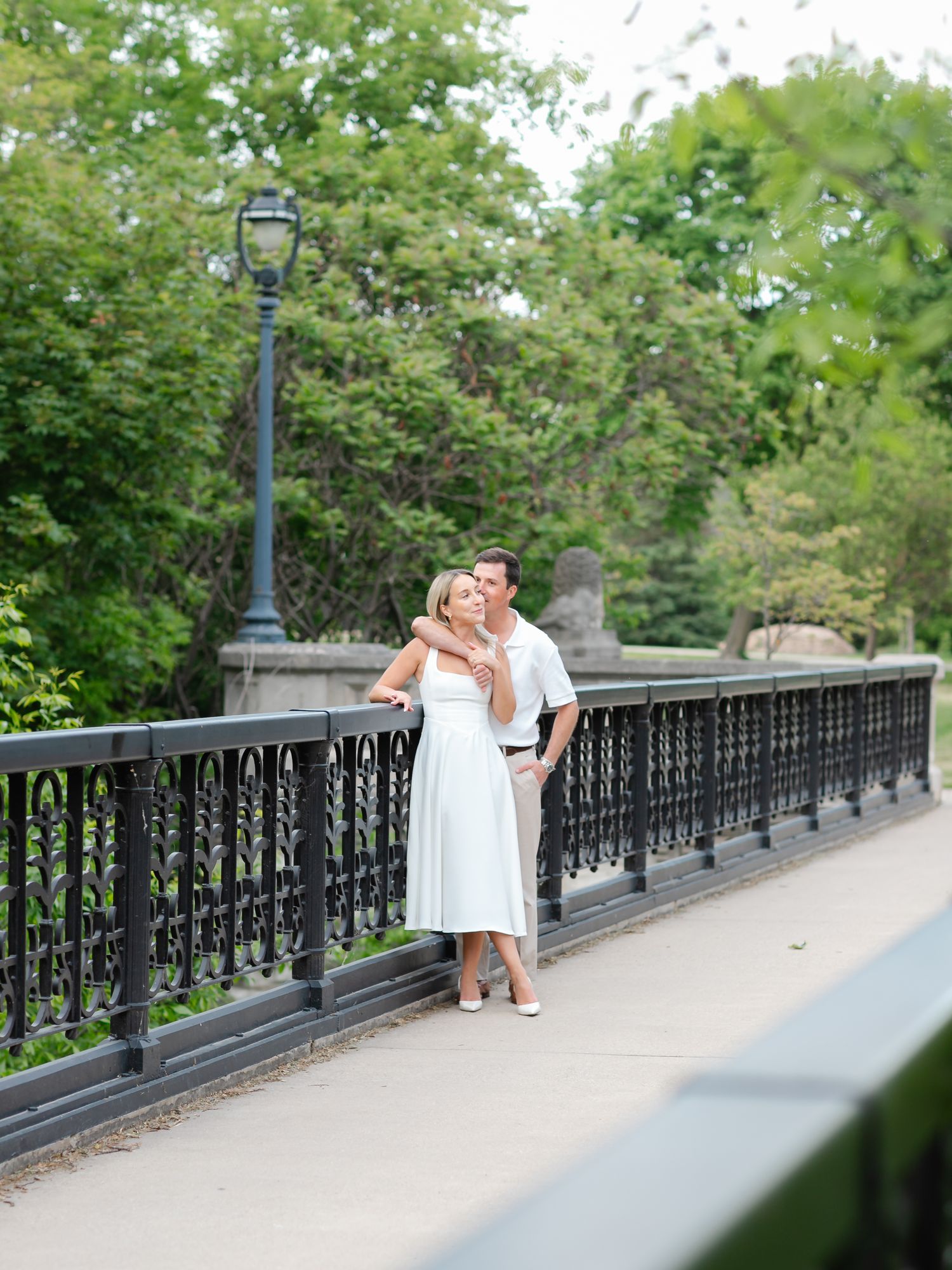 Couple in white outfits embracing on a decorative iron bridge in a lush green park setting.