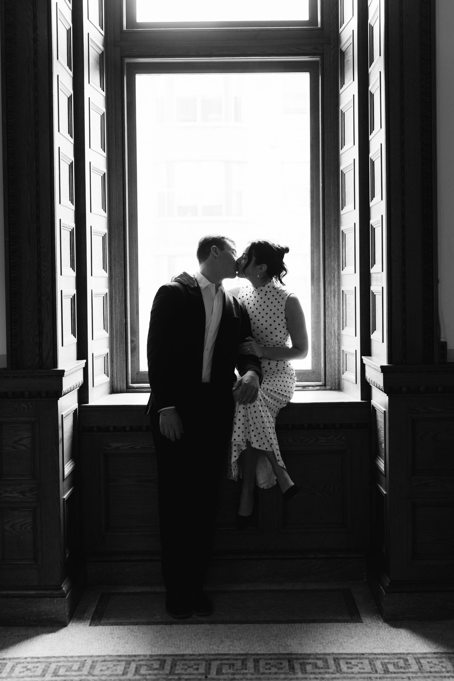 Black and white photo of a couple sharing a kiss by a grand window in an elegant historic building.