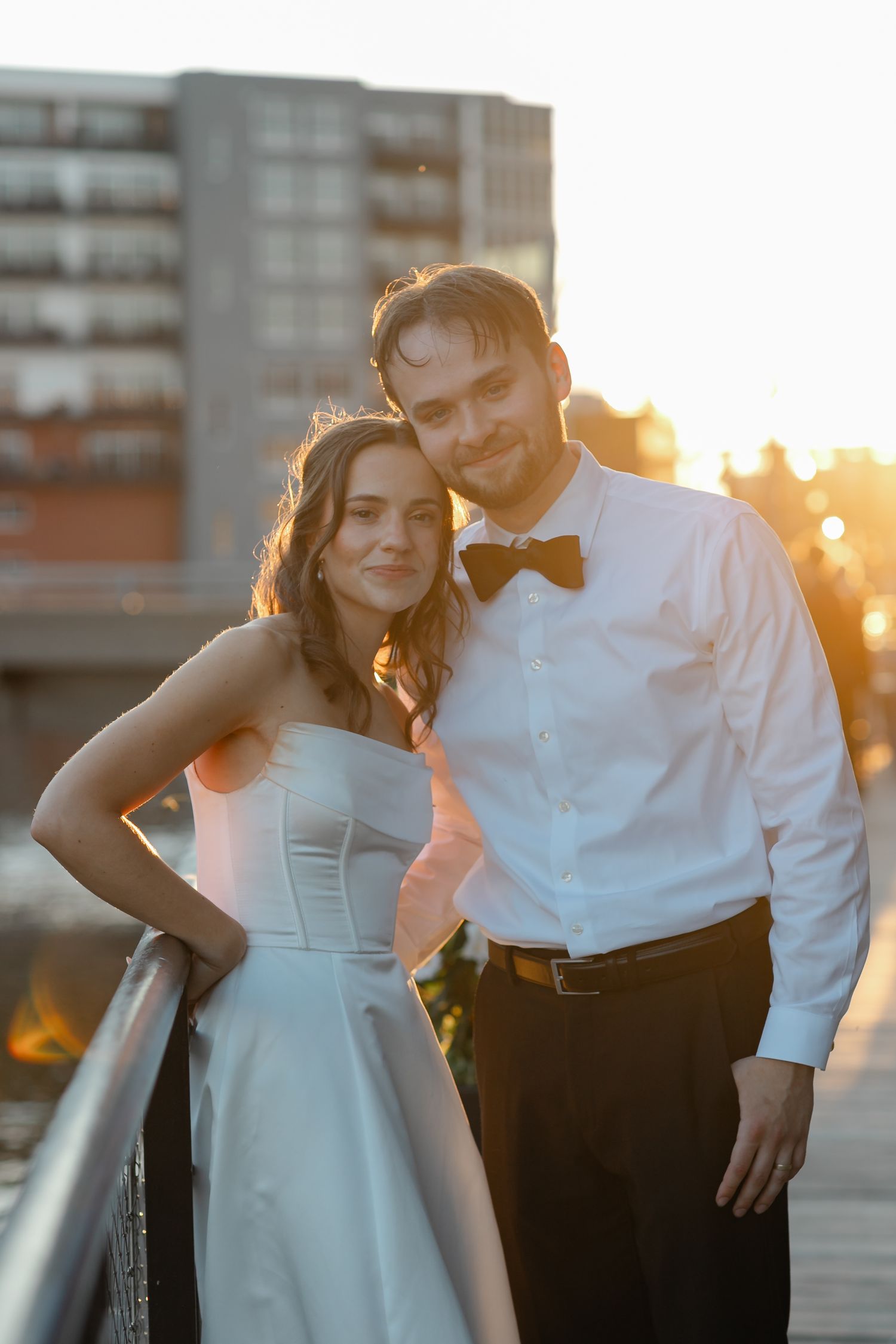 Bride and groom posing together at sunset on a rooftop, wearing wedding attire with a city skyline backdrop.