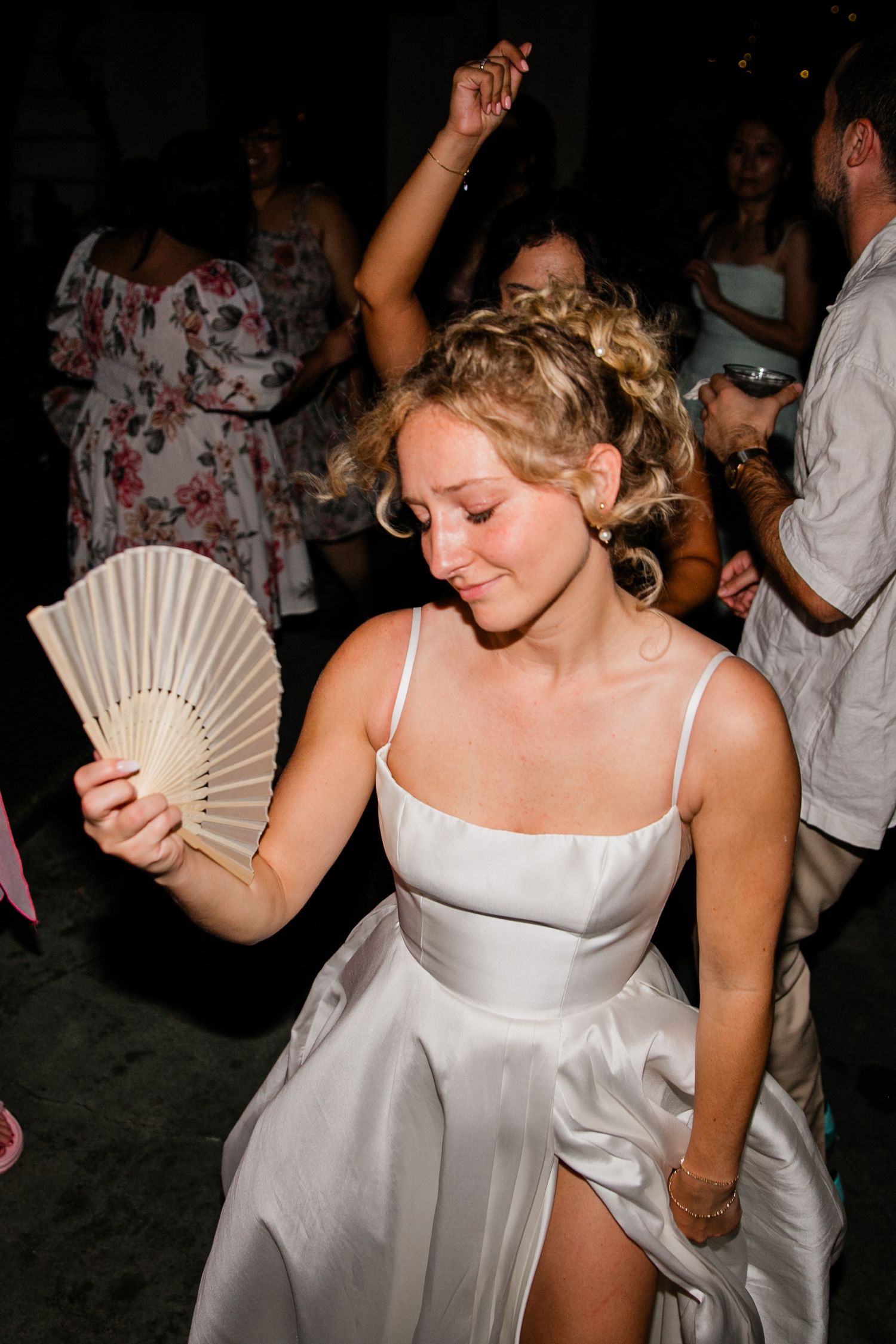 A woman in white dress cools herself with a paper fan while dancing at a wedding reception.