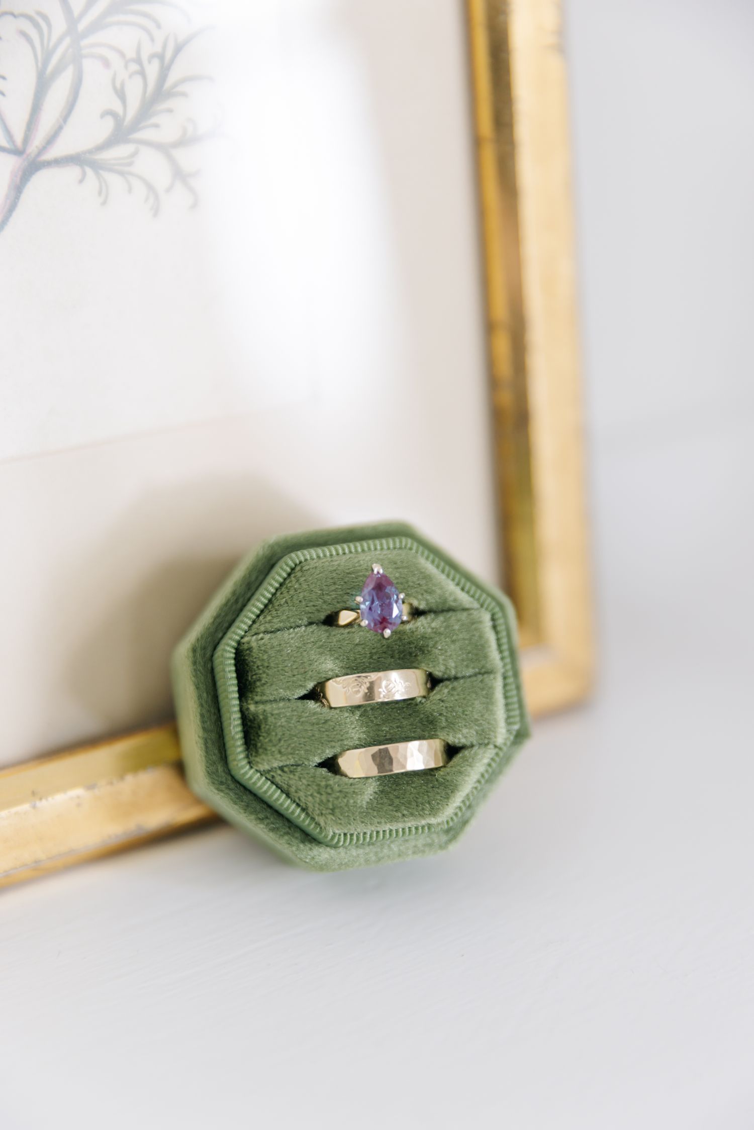 Green velvet octagonal ring box holding two gold wedding bands and a pear-shaped purple sapphire engagement ring.