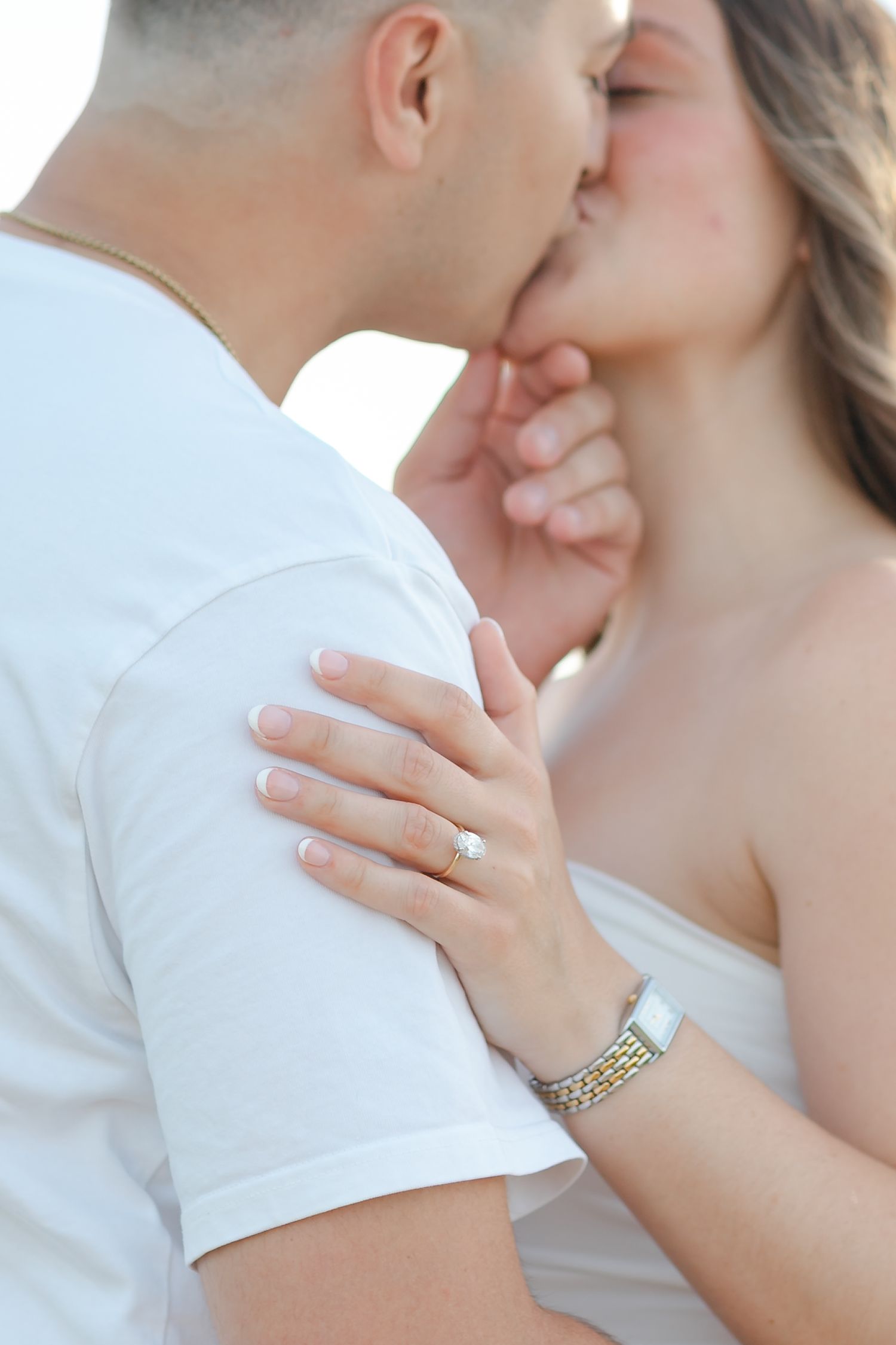 Couple sharing a romantic kiss, woman wearing engagement ring and bracelet, man in white t-shirt.