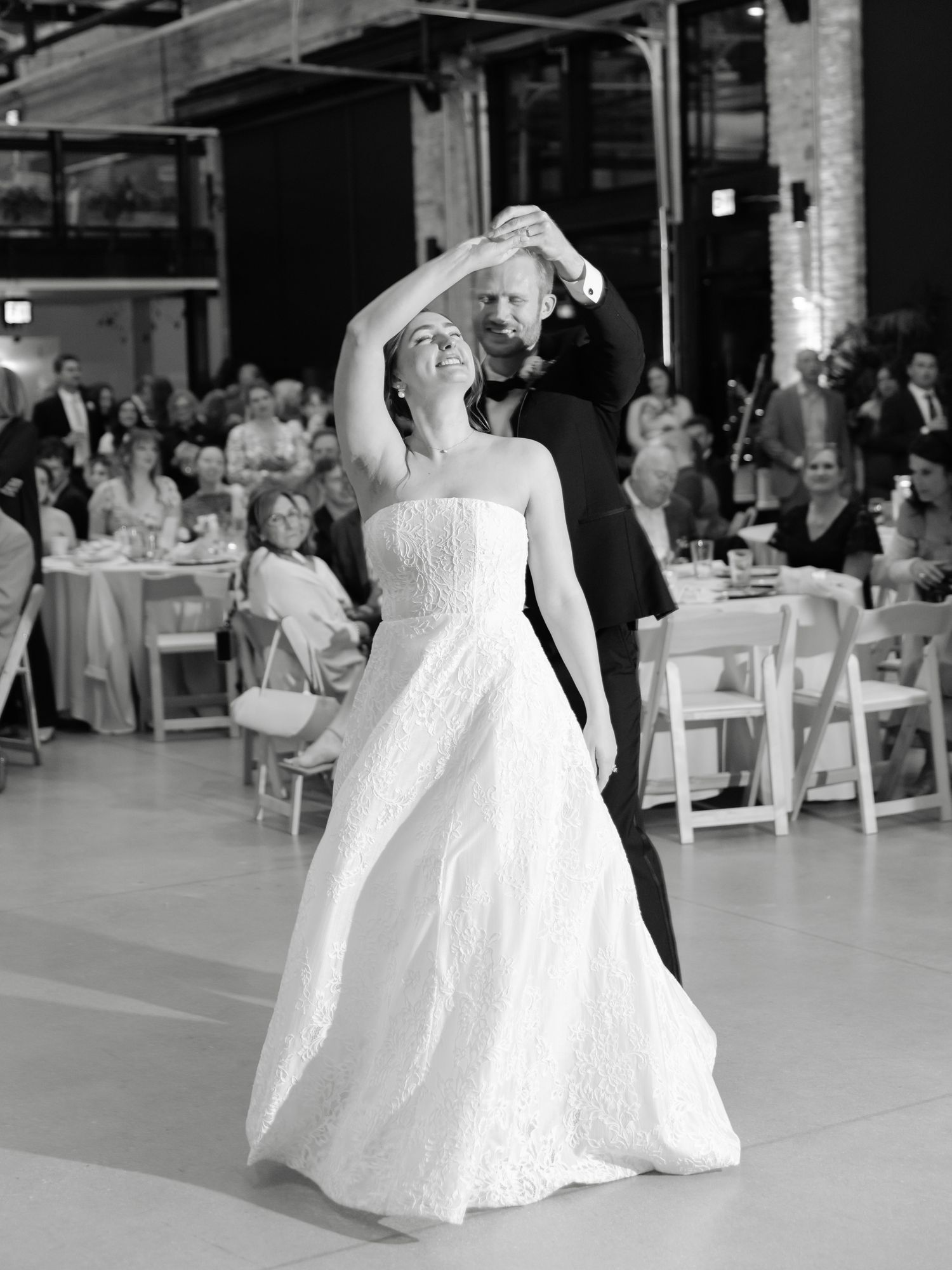 Bride and groom share their first dance at wedding reception, bride in strapless gown being twirled by groom.