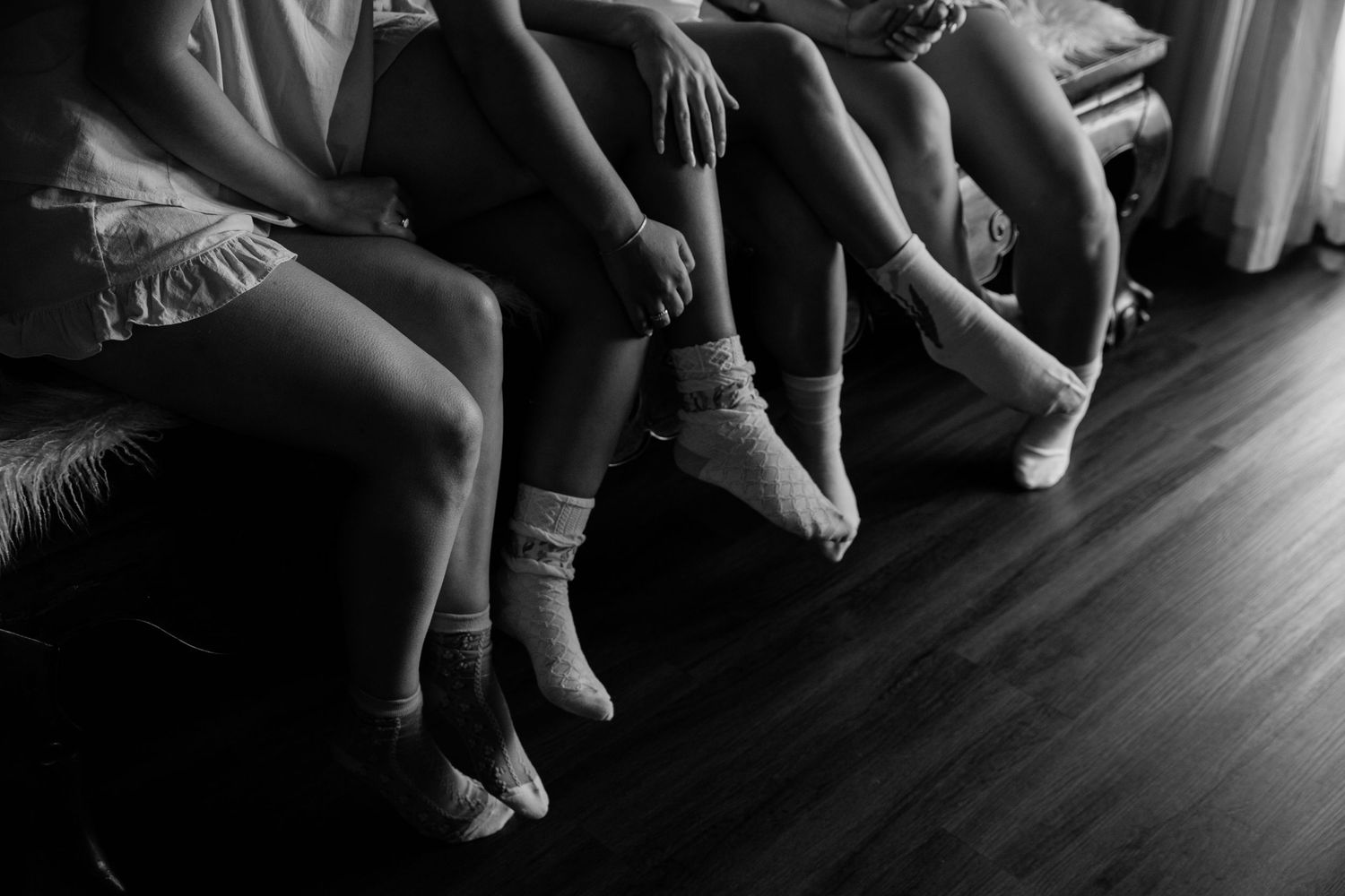 Black and white image of bare feet with anklets dangling over a hardwood floor.