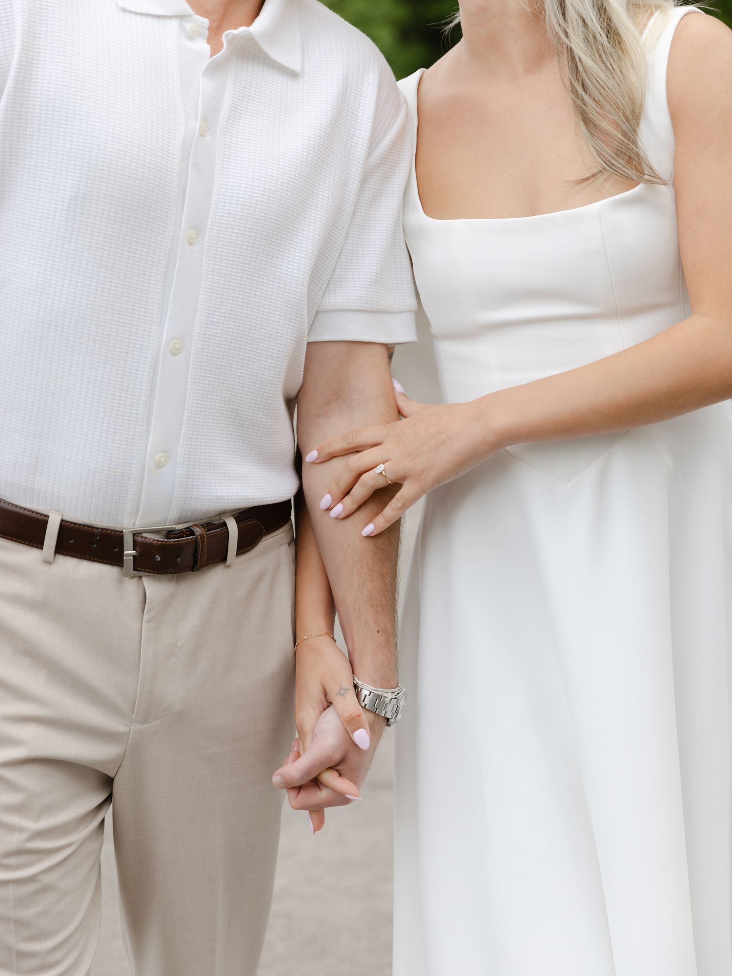 Close-up of engaged couple holding hands, woman wearing diamond ring, both dressed in white and beige.