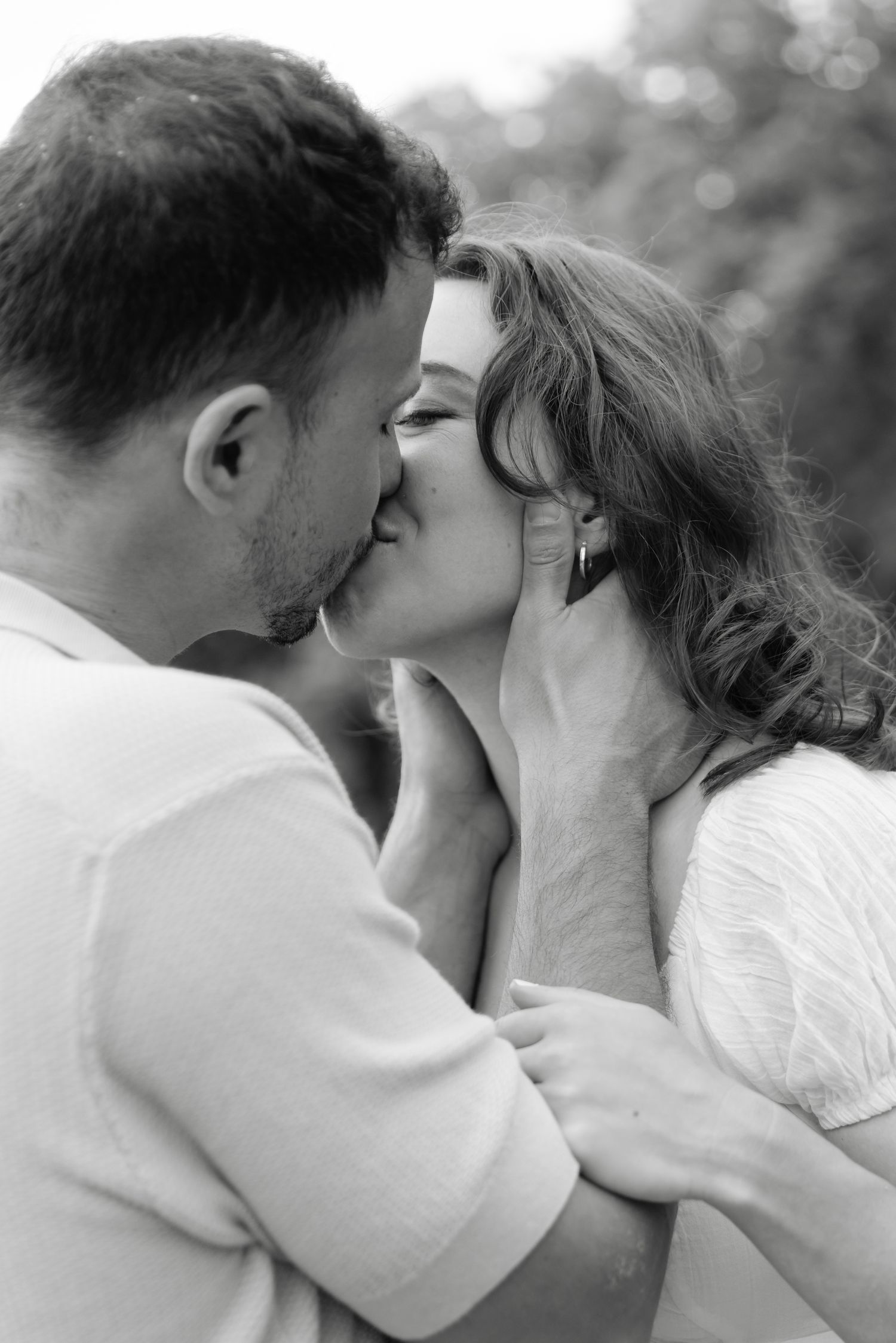 Black and white photo of a romantic couple sharing a tender kiss outdoors, man gently holding womans face.