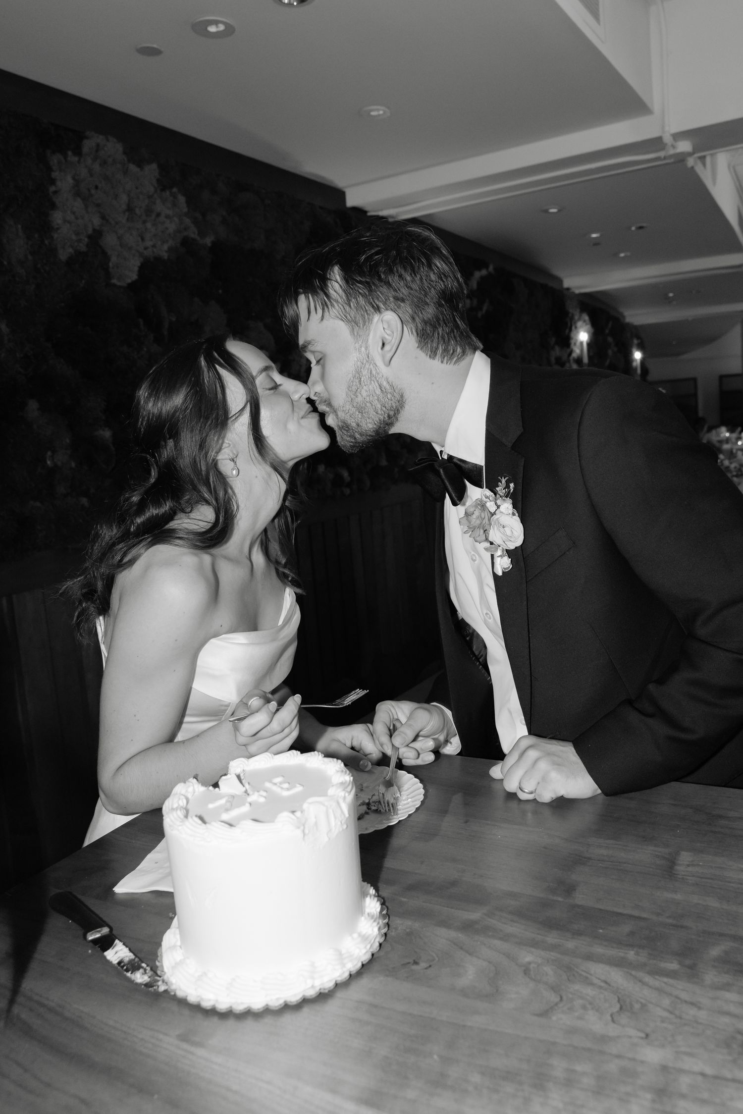 Black and white photo of a bride and groom sharing a kiss while cutting their wedding cake at the reception.