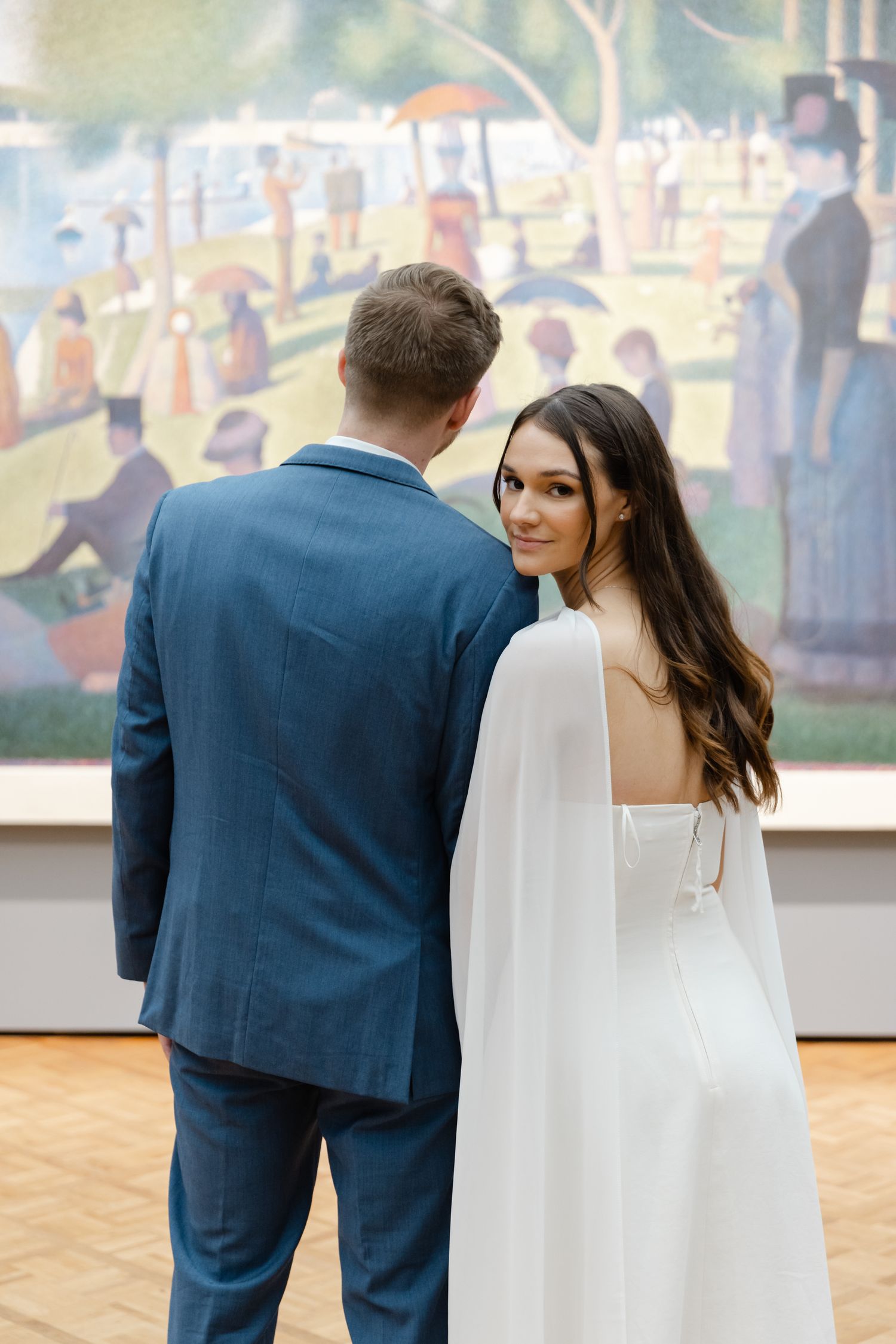 Bride in white cape gown and groom in blue suit viewing a famous painting at an art museum wedding.