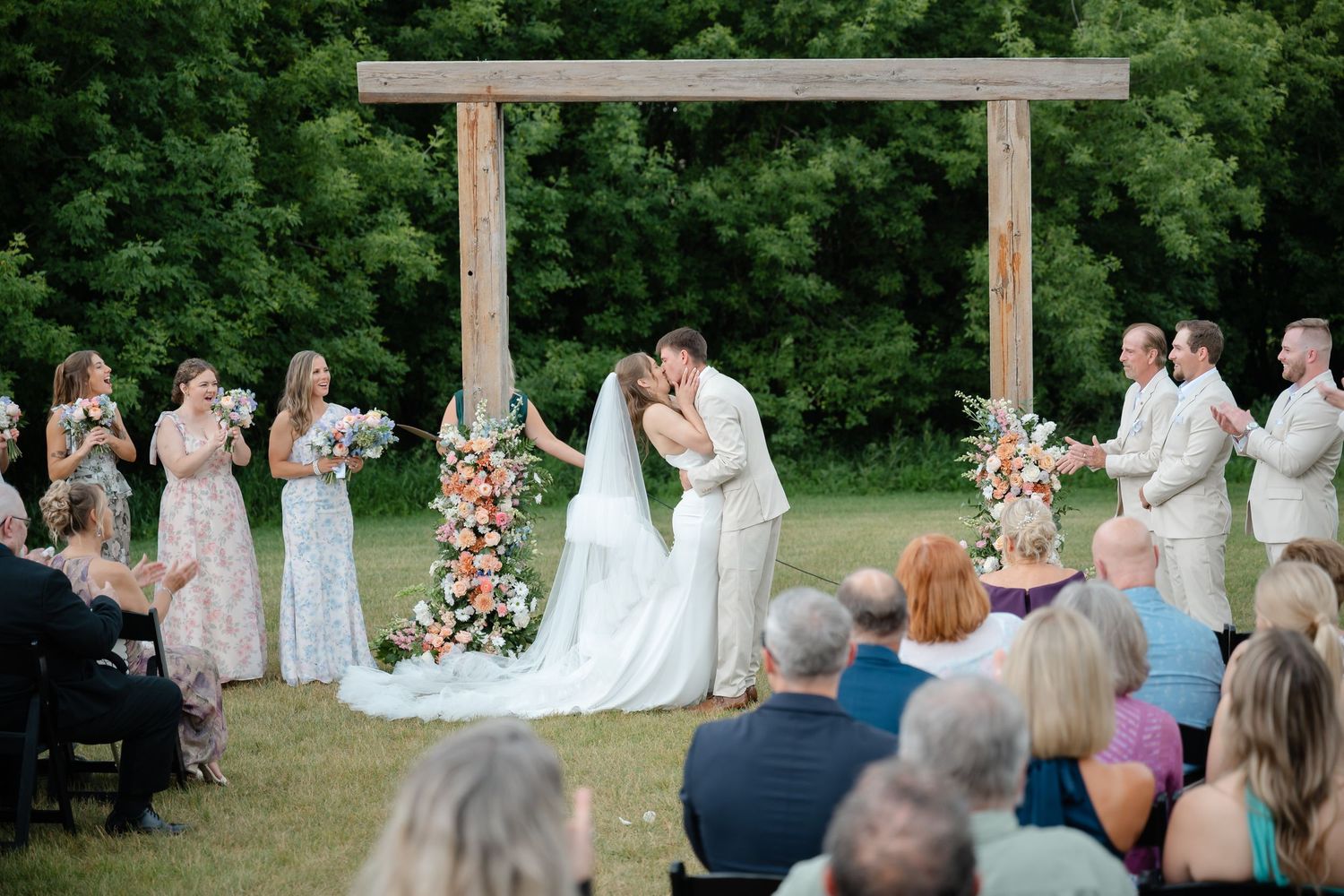 Bride and groom share first kiss under rustic wooden arch during outdoor wedding ceremony with guests and bridal party.