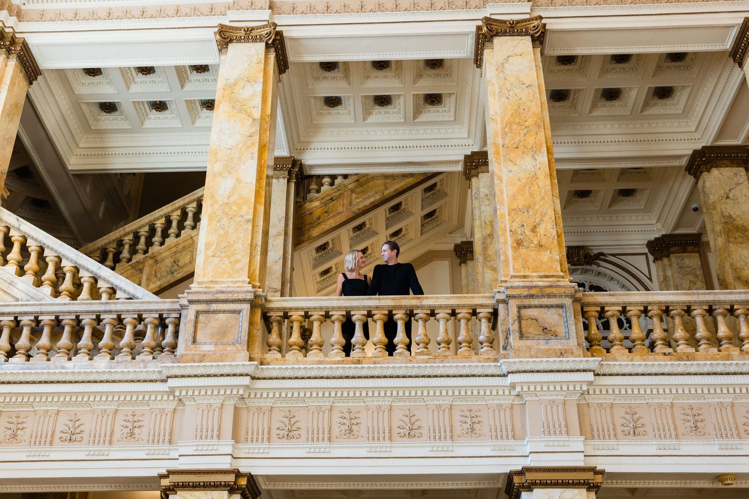 Couple stands on ornate marble balcony with golden columns in elegant historic building interior.