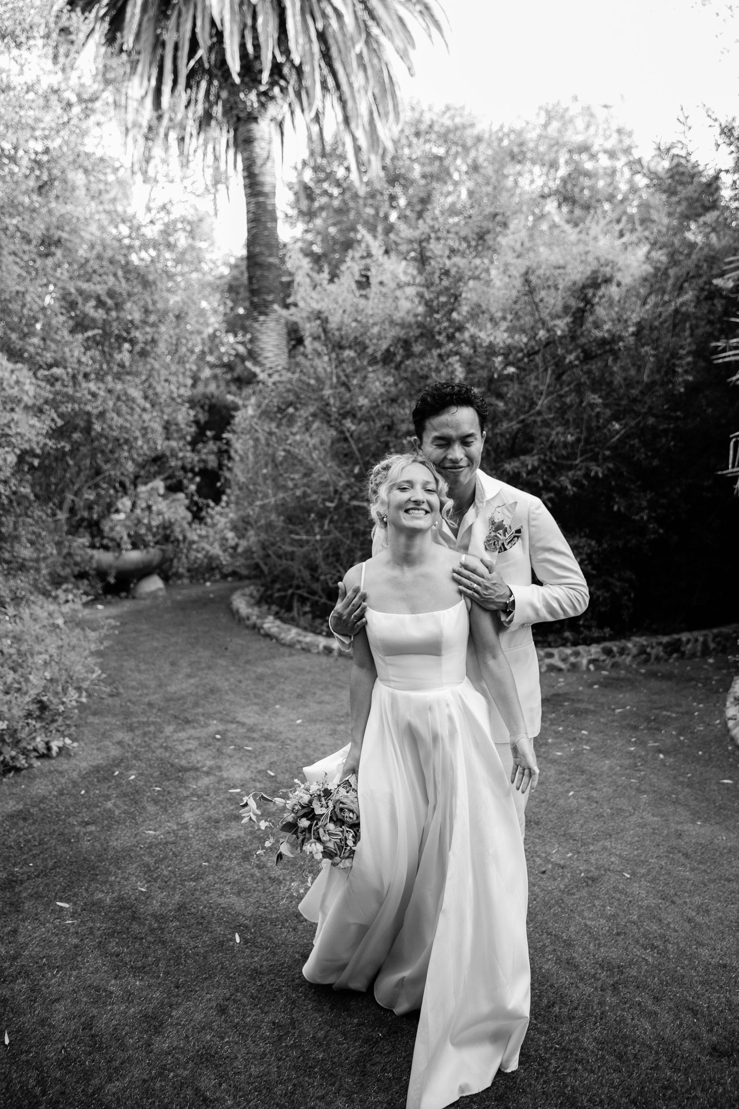 A black and white wedding photo of a couple sharing a joyful moment in a garden with palm trees.