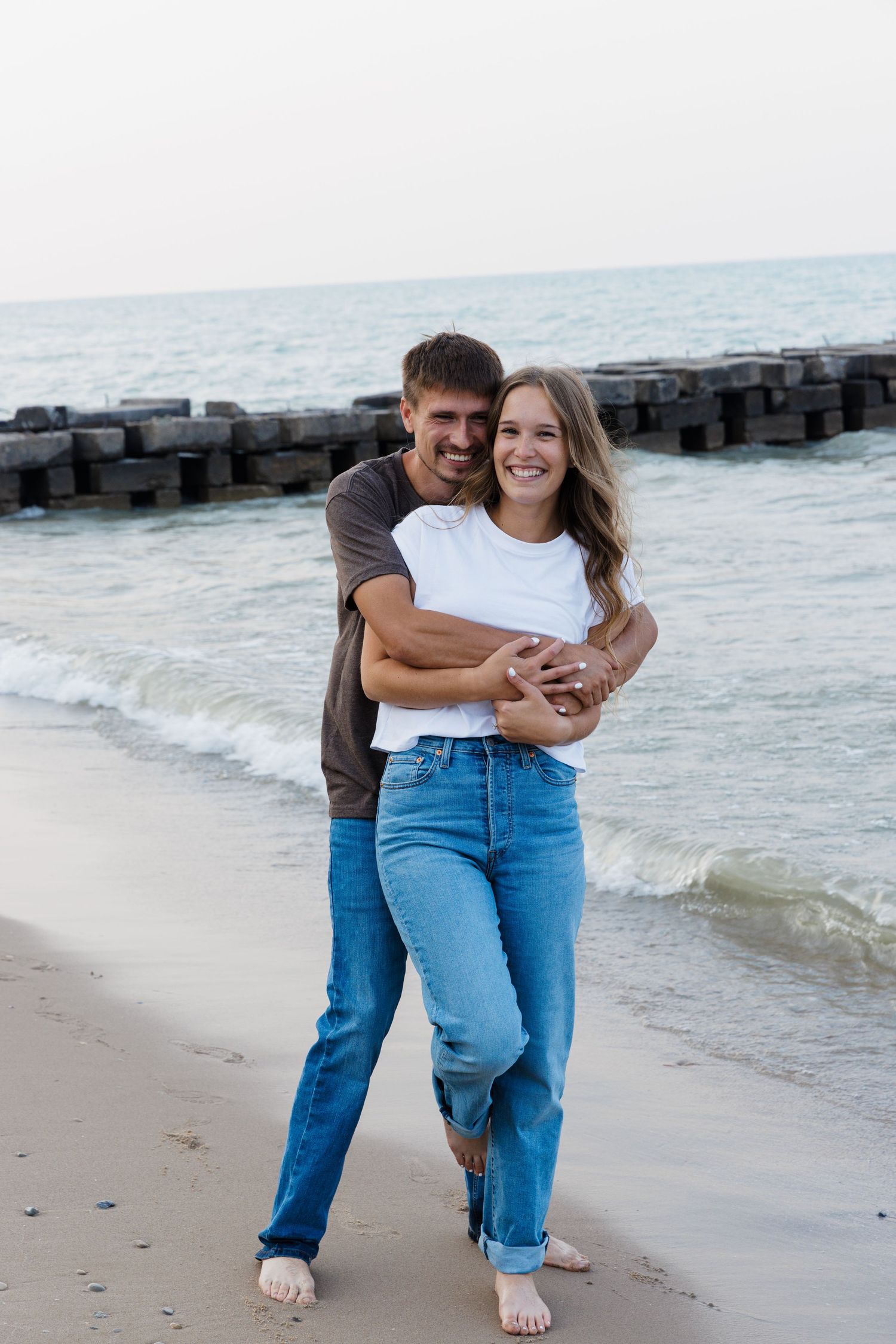 Happy couple embracing on a sandy beach, smiling with ocean waves and wooden pier in the background.