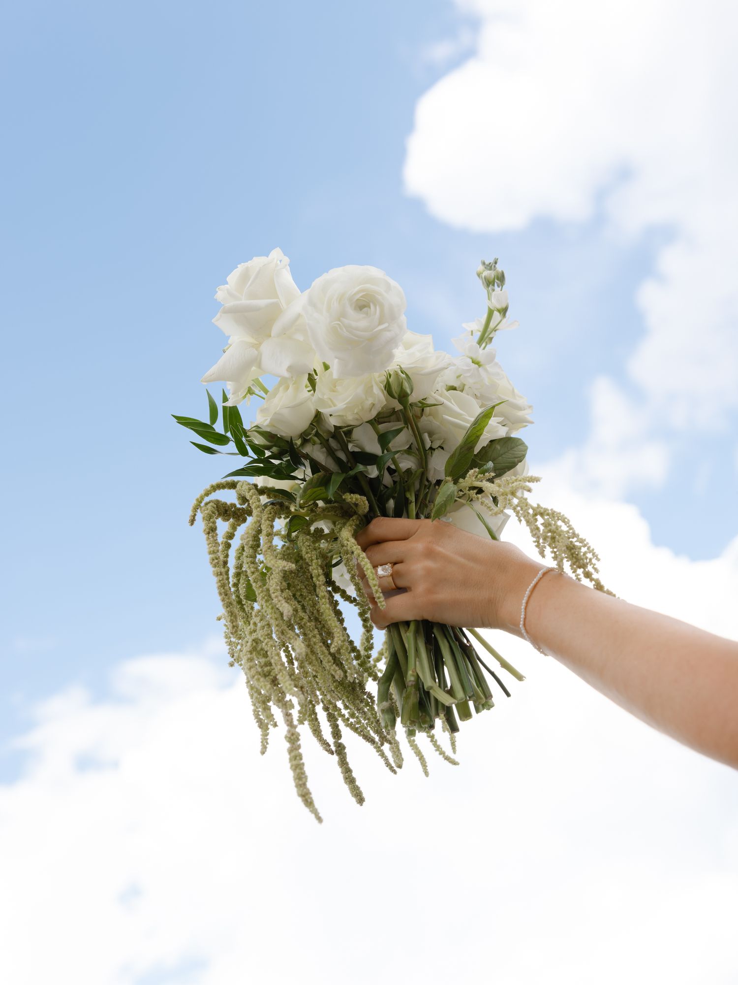 A hand holds a white bridal bouquet of roses and greenery against a bright blue sky with soft clouds.