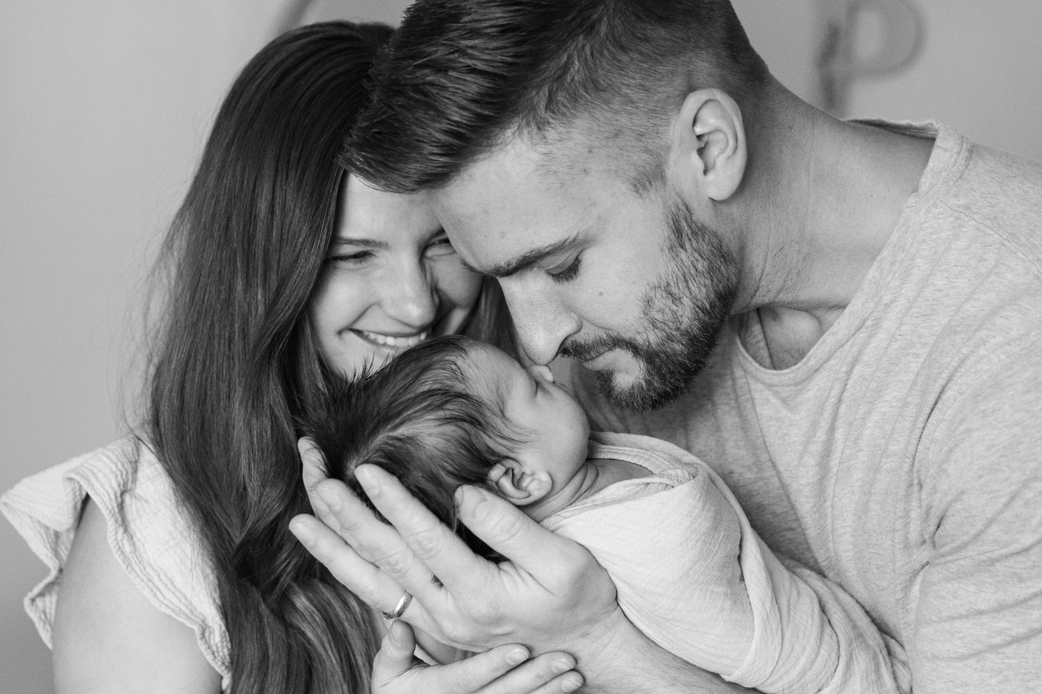 Black and white photo of loving parents tenderly kissing their newborn baby together in a heartwarming family portrait.