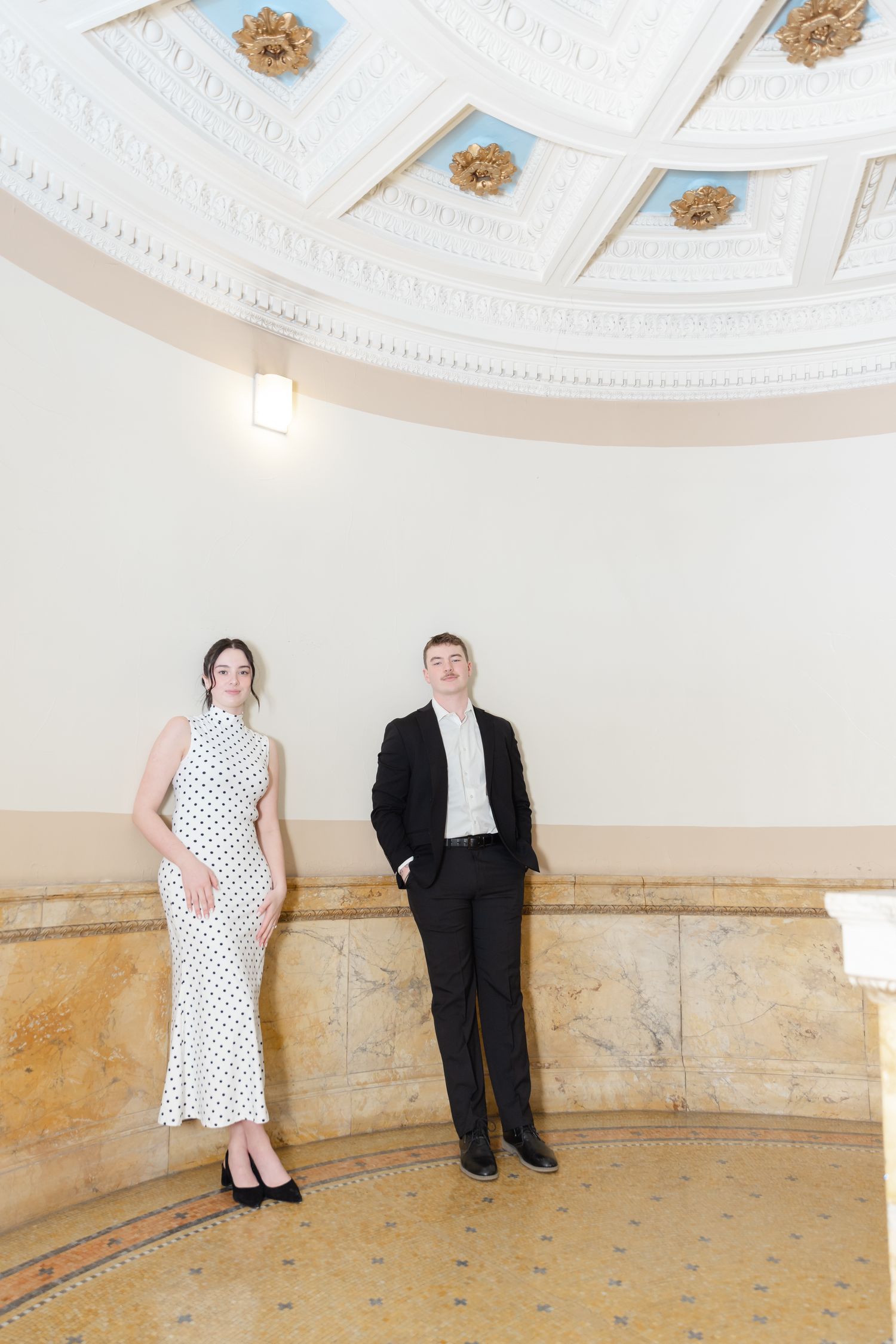 Elegant couple in formal attire posing in a grand rotunda with ornate ceiling details and marble walls.
