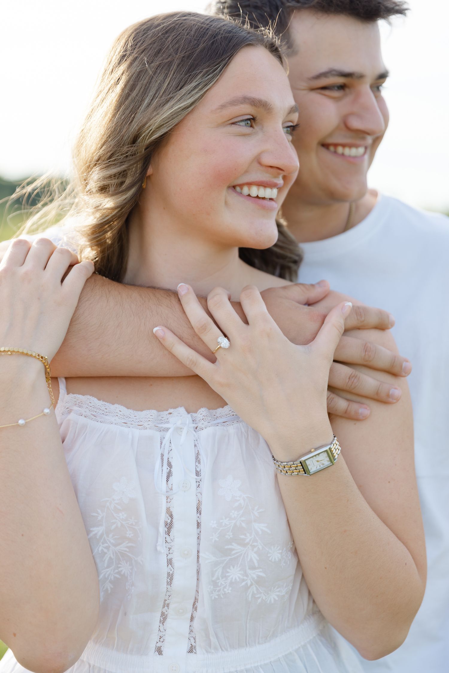 Happy engaged couple embracing outdoors, woman in white dress showing diamond ring, wearing gold bracelet and watch.