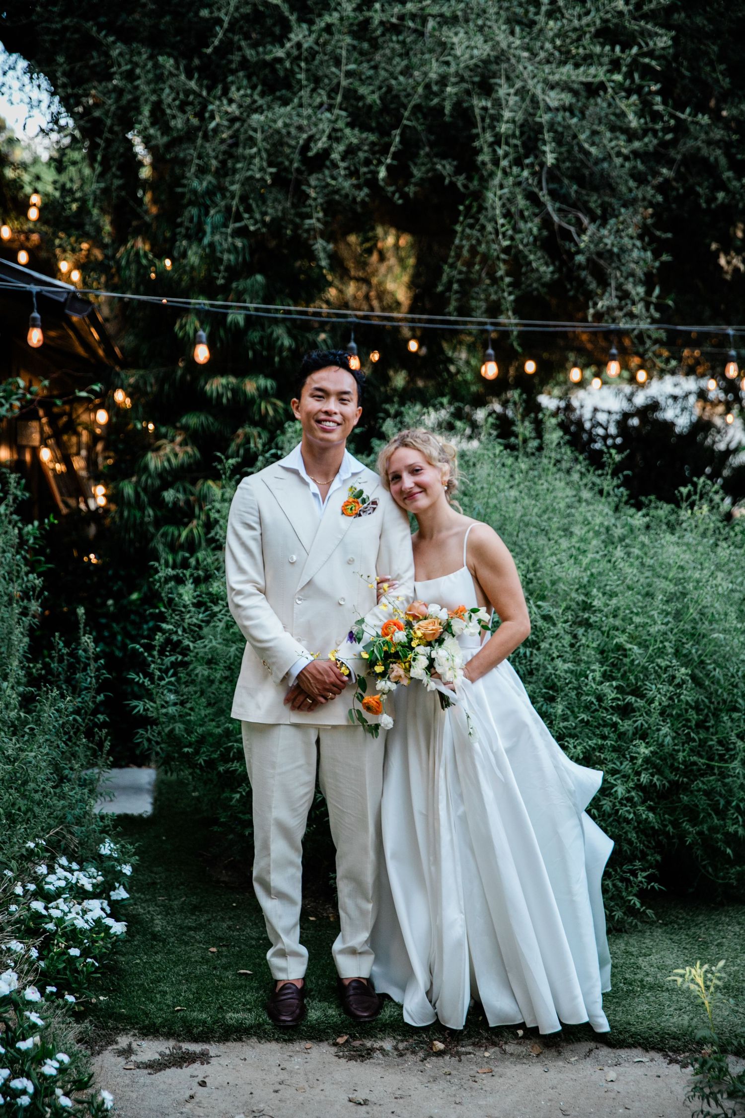 Wedding couple poses in garden setting with string lights and natural greenery backdrop.
