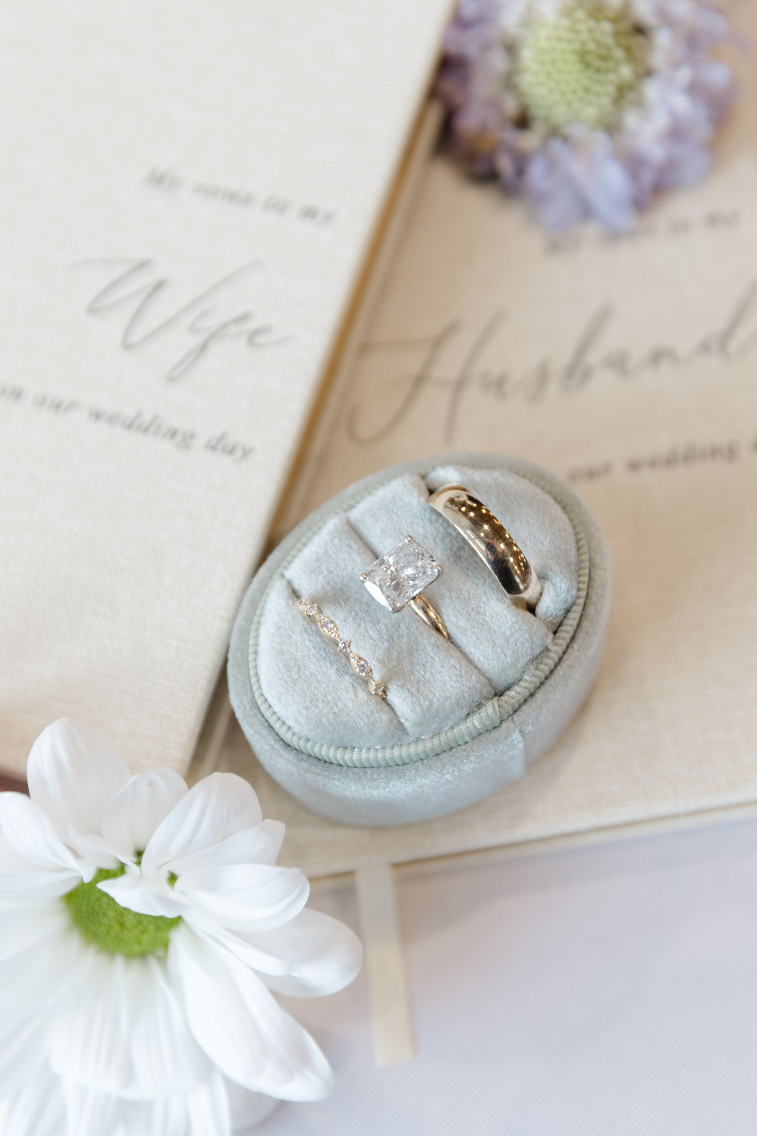 Wedding rings in velvet box with vow books labeled Wife and Husband, accented by white daisy flowers.