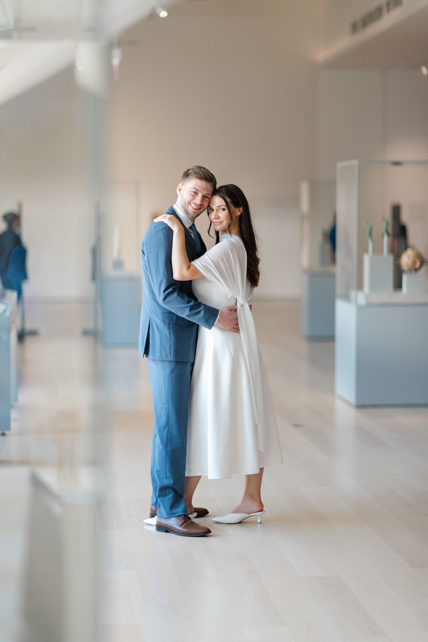 Newlyweds embracing in a bright museum gallery, groom in blue suit and bride in elegant white dress.
