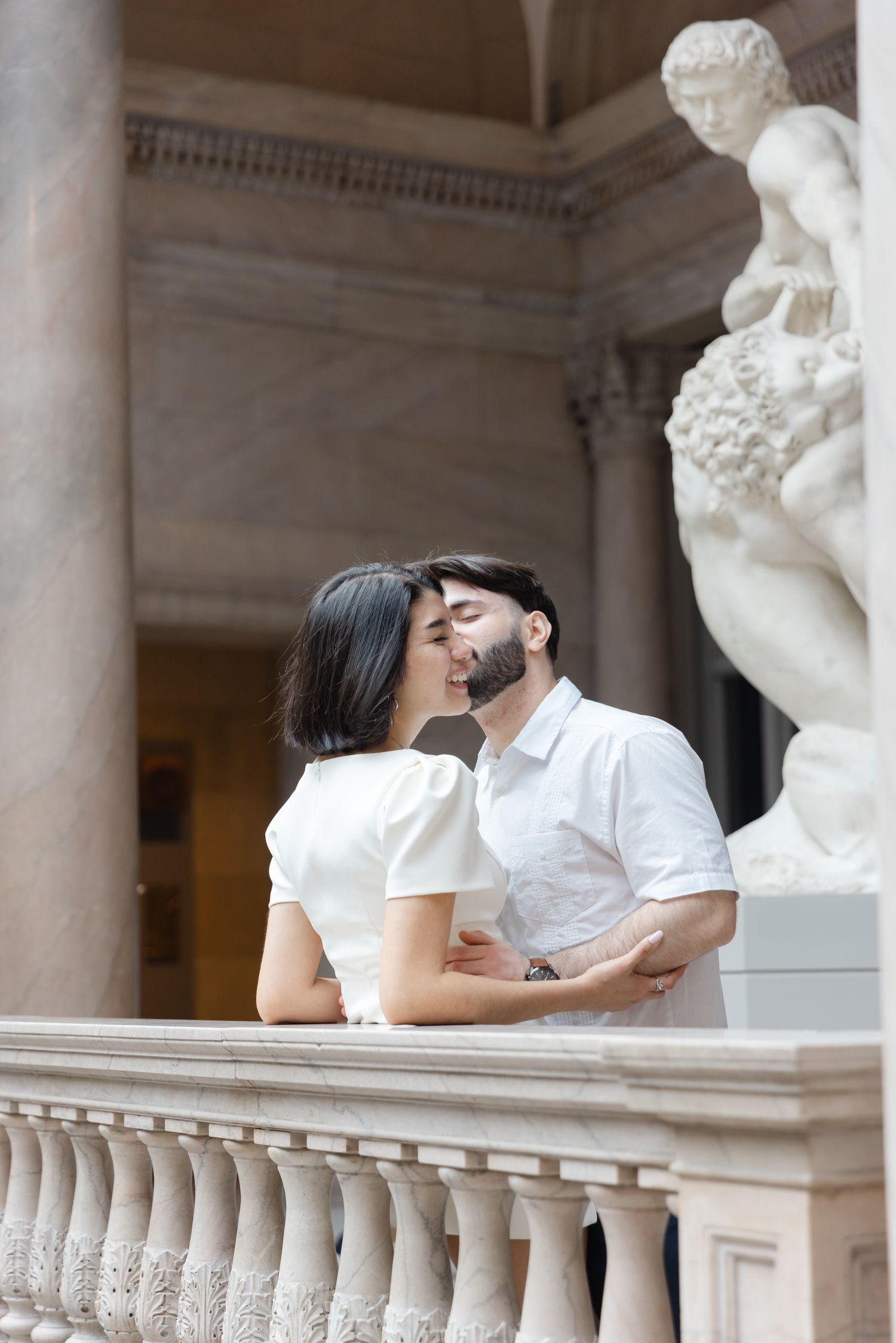Couple sharing a kiss near ornate stone balustrade with classical sculpture in elegant museum gallery.