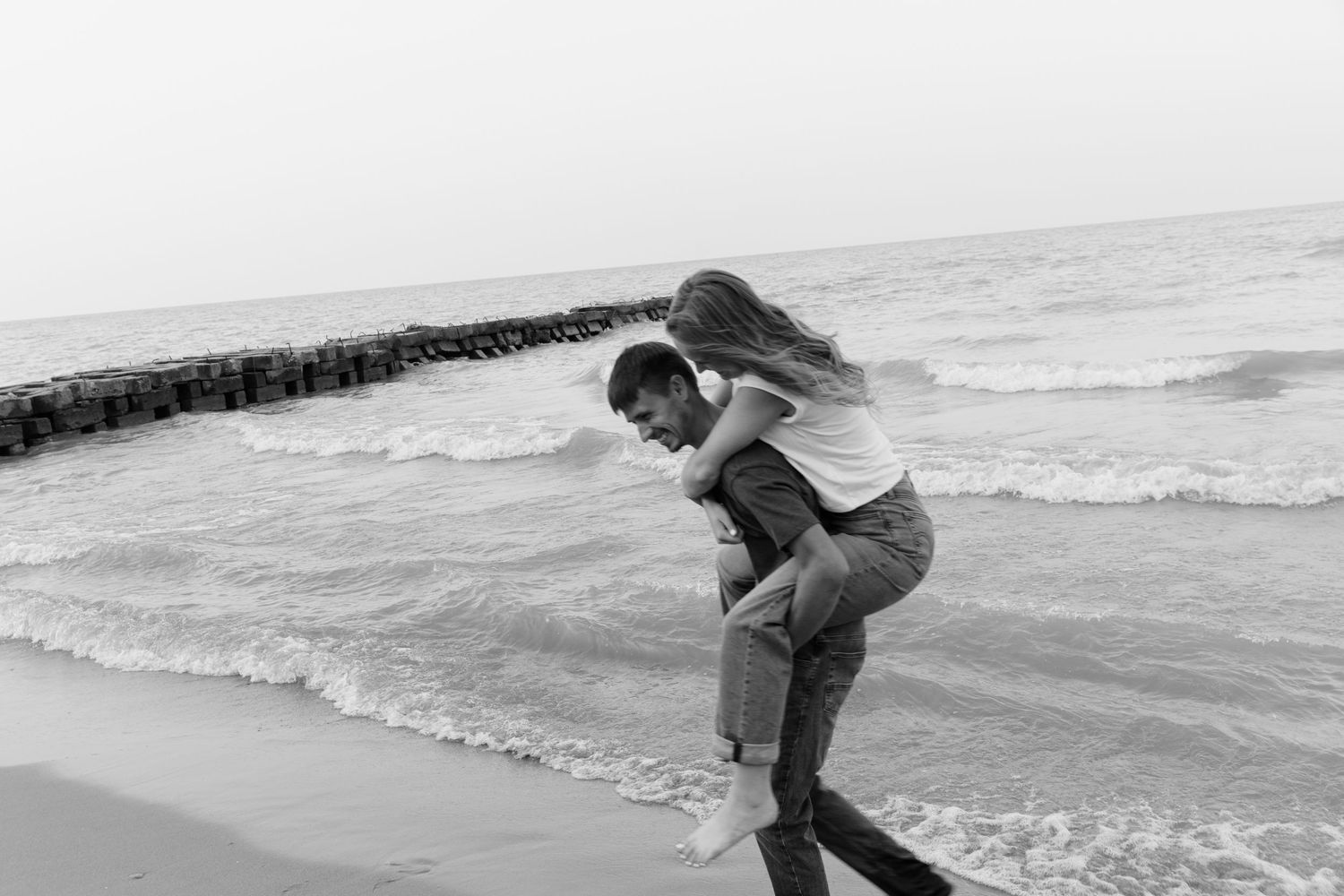 Black and white photo of a couple playing on the beach, man giving woman a piggyback ride by the ocean waves.