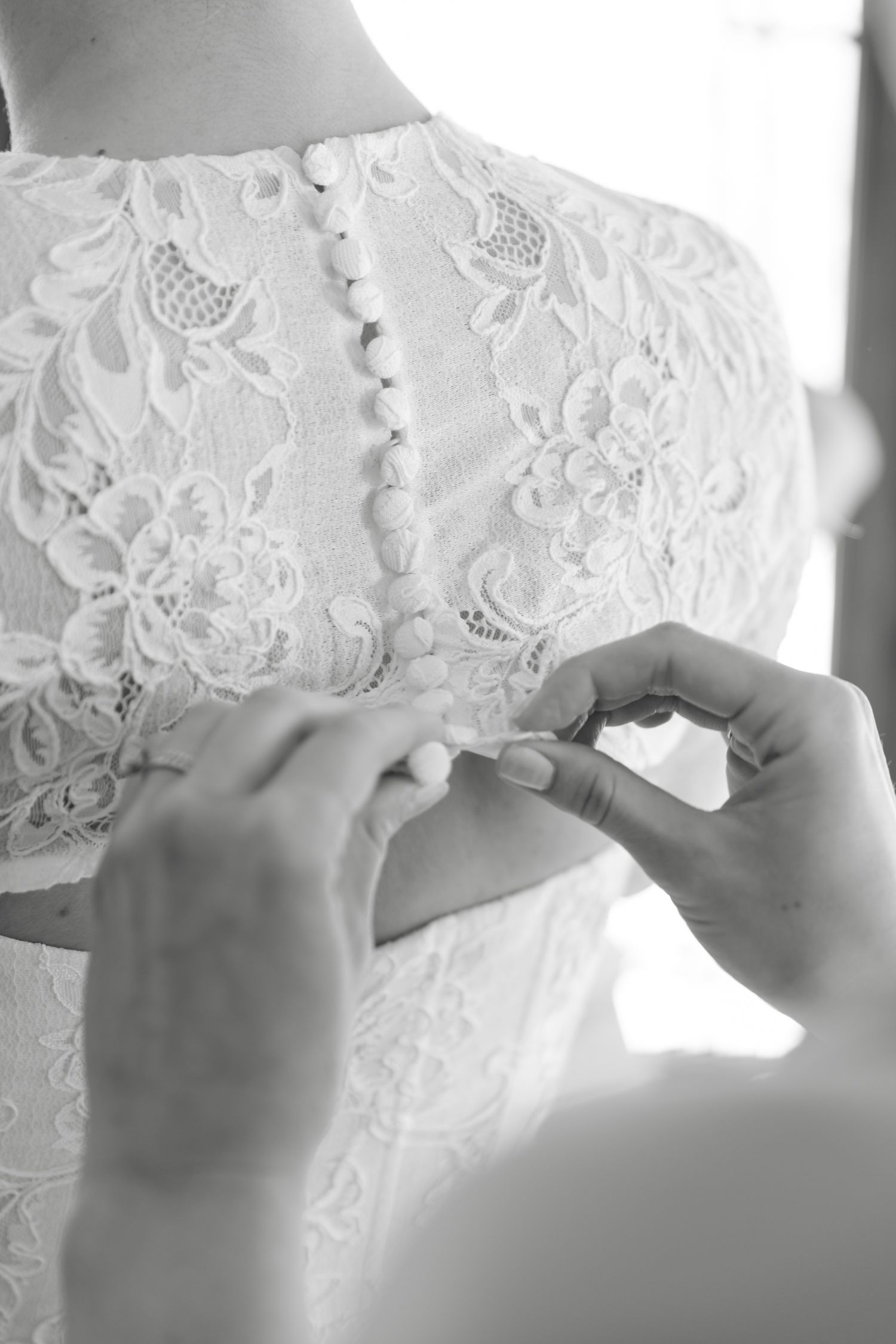 Black and white photo of hands buttoning the back of a lace wedding dress with pearl buttons.