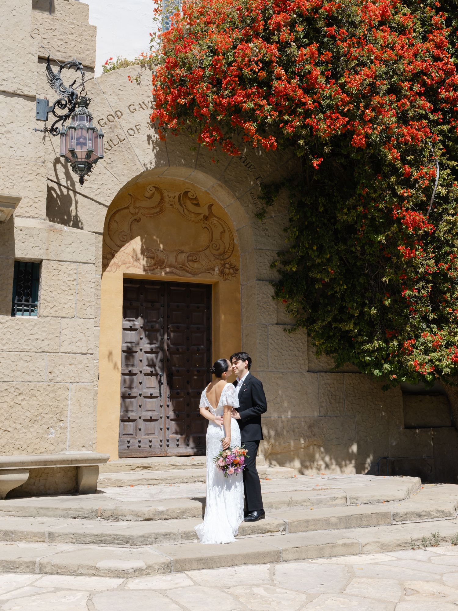 Bride and groom kissing under a stone arch adorned with vibrant red bougainvillea at an elegant outdoor wedding venue.
