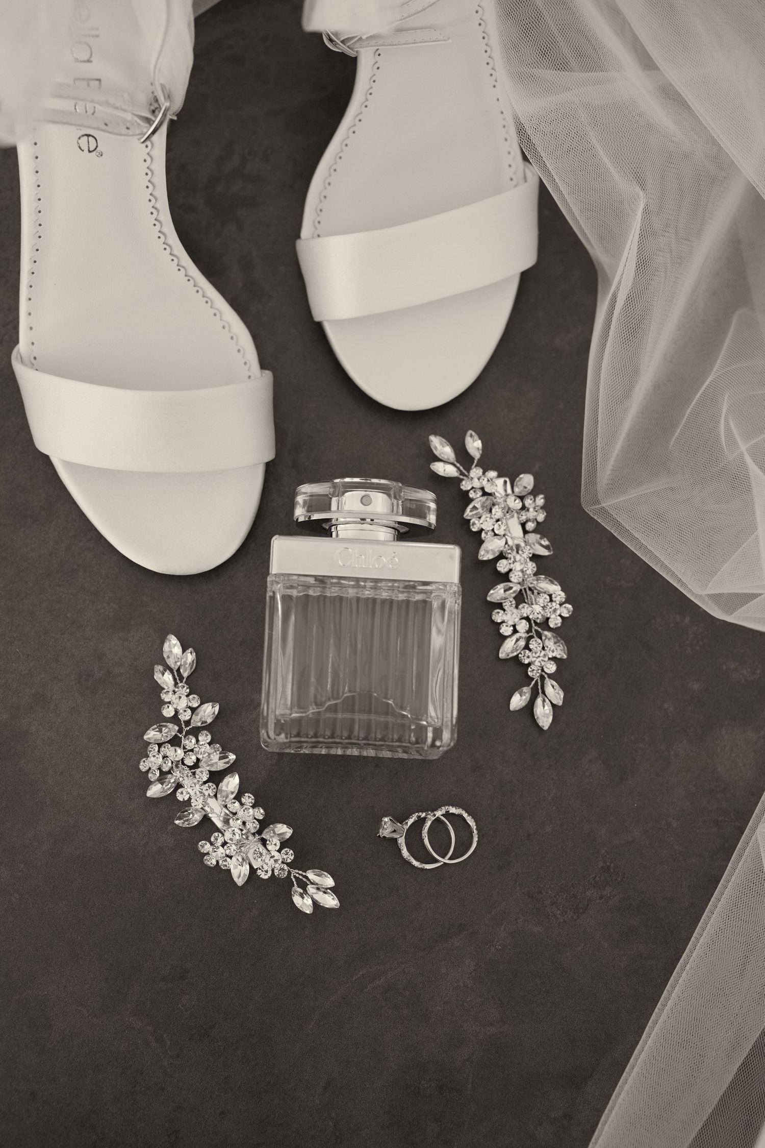 Wedding accessories including white sandals, perfume bottle, floral hair pieces, and wedding rings arranged on dark surface.