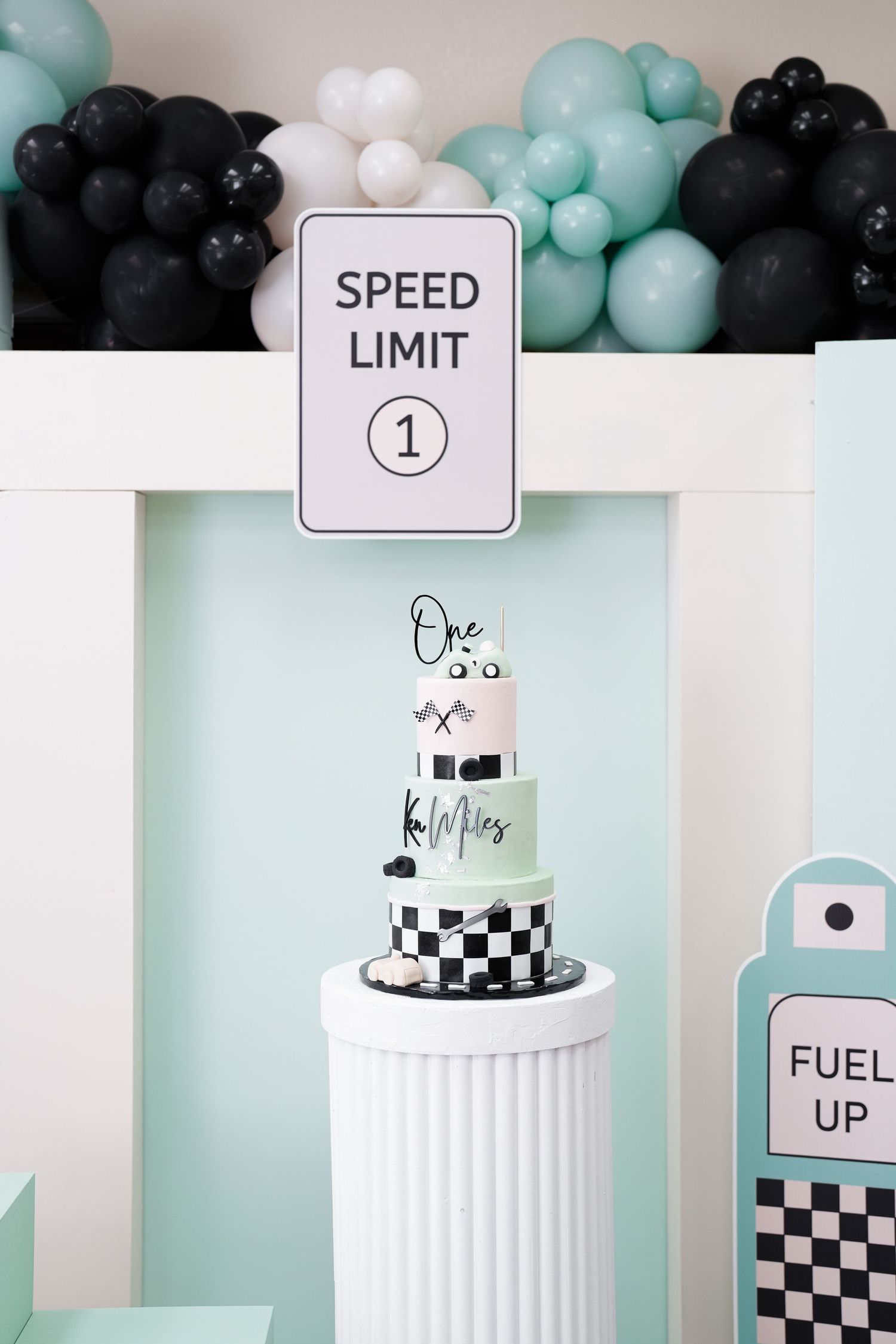 A racing-themed birthday cake display with black and mint green balloons, speed limit sign, and checkered pattern decorations.